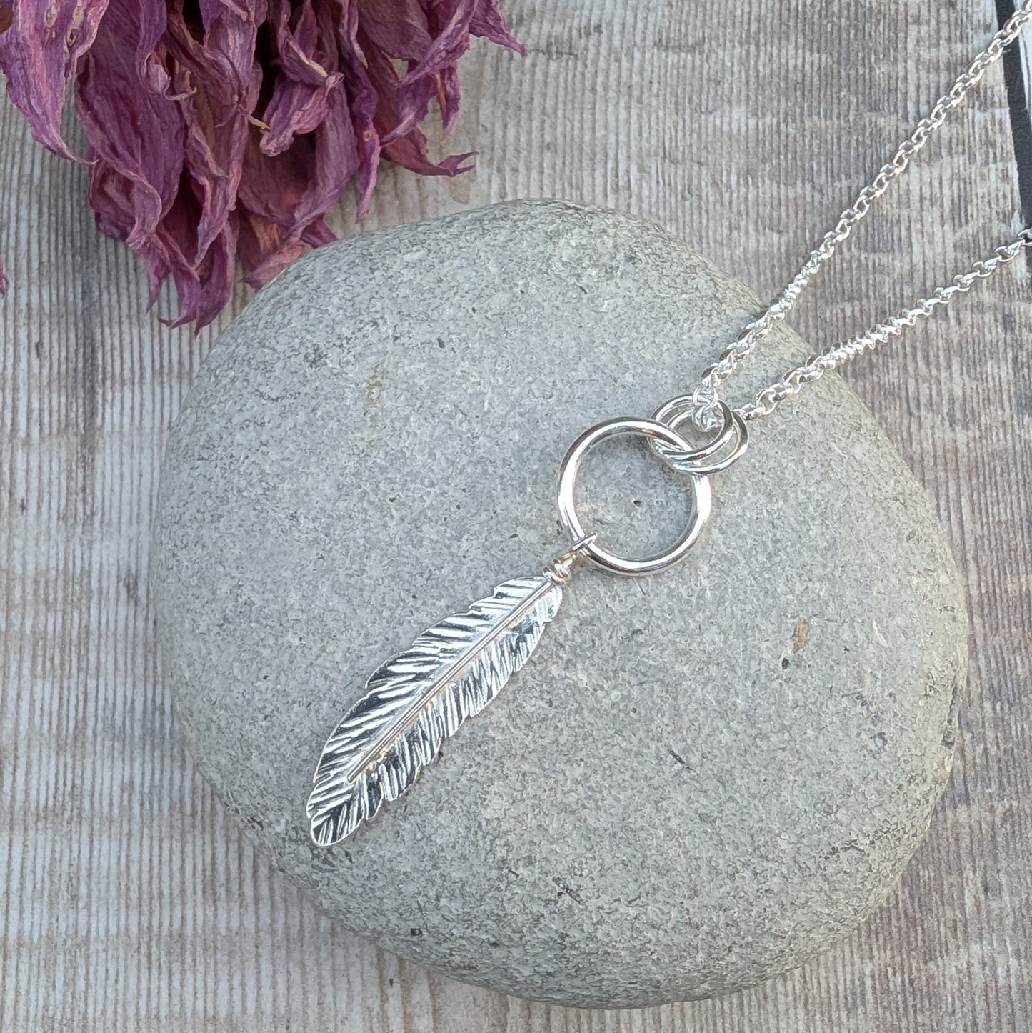 A handmade sterling silver necklace featuring a detailed feather pendant hanging from a circular silver ring, displayed on a smooth grey stone. The necklace chain extends out of the frame, and dried purple flowers are in the background.