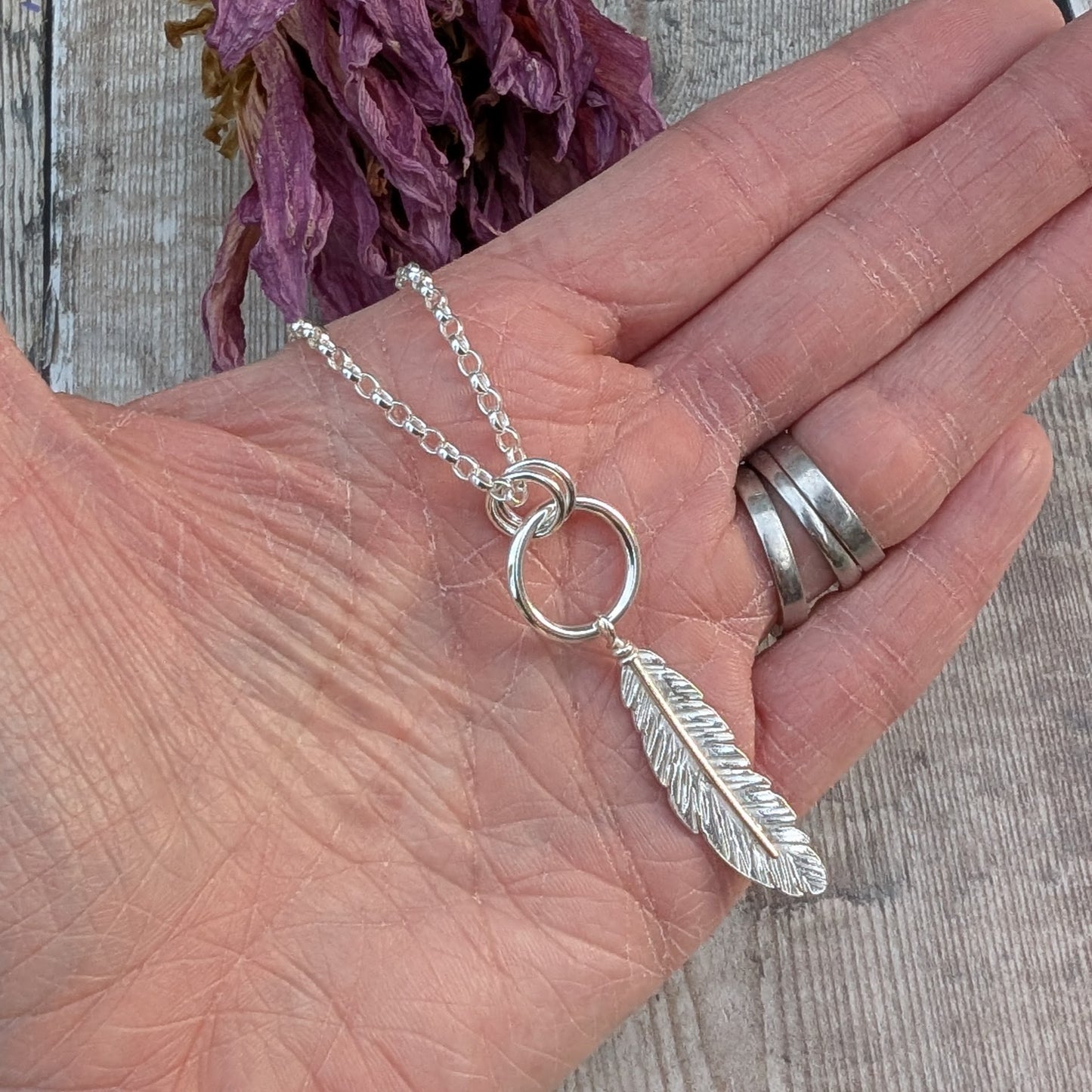 The silver feather and circle necklace resting on an open palm, showing its scale. The intricate feather pendant hangs below the interlocking rings, with the silver chain draping over the fingers. The background is a wooden surface with dried purple flowers.