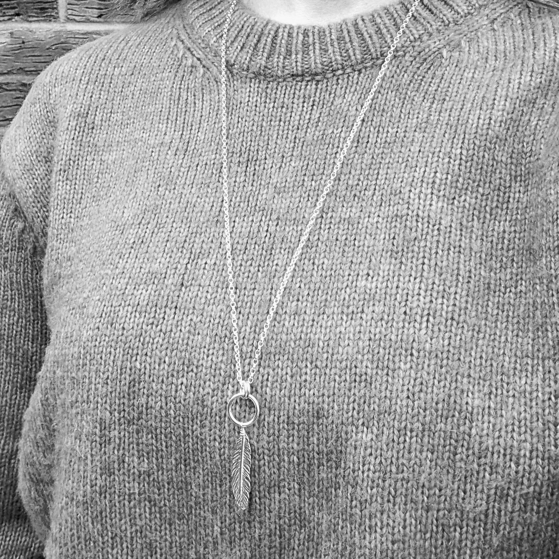 A black-and-white image of a person wearing the long silver feather and circle necklace over a textured knit sweater. The pendant hangs at mid-chest level, showing its length and how it sits when worn.
