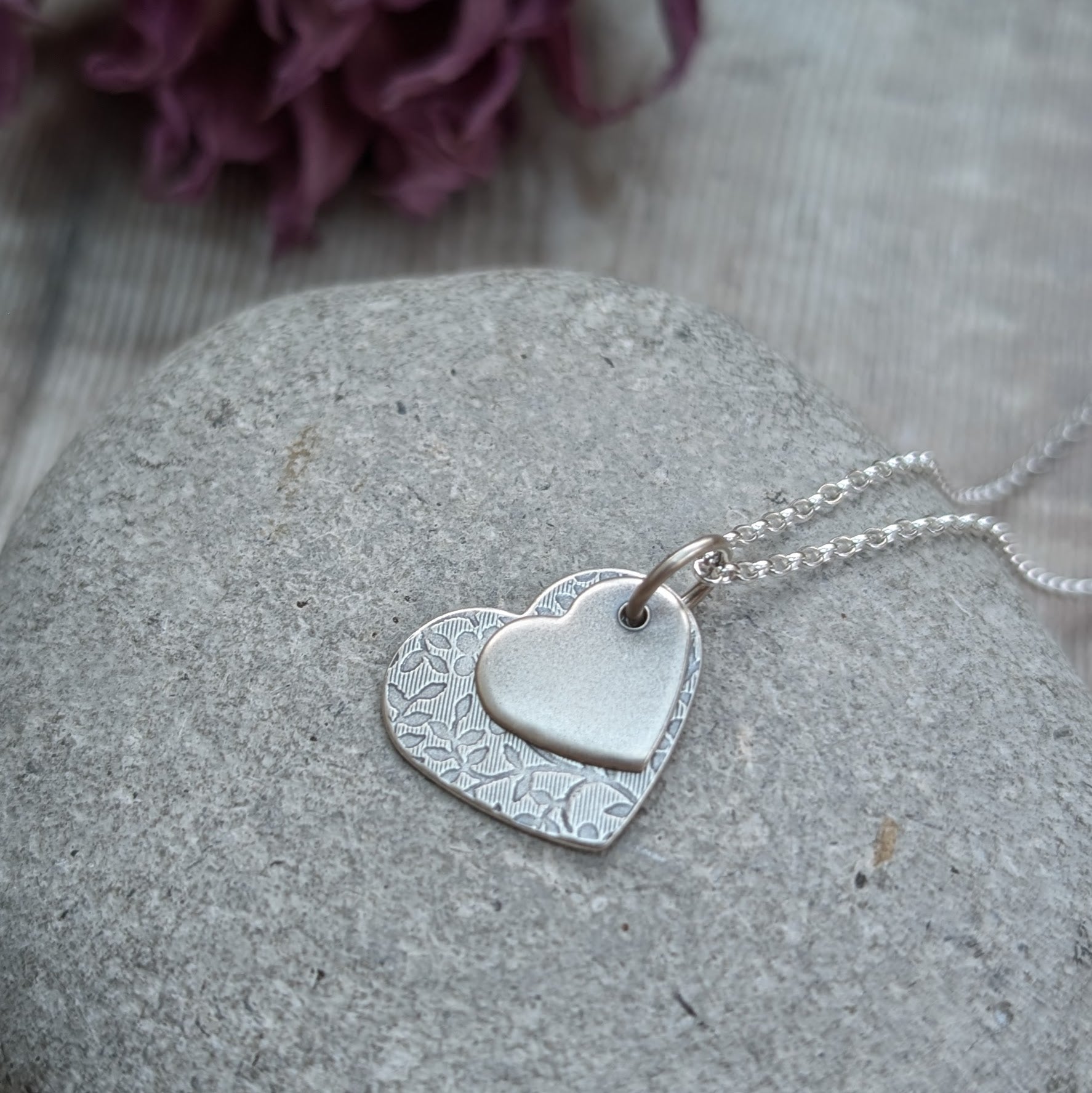 Pendant is a flat silver heart shape with detailed stamped floral pattern, overlapped by a smaller flat silver heart shape with softly brushed finish. The hearts have been oxidised to enhance the floral details. Suspended from a silver chain via a small circle link. 