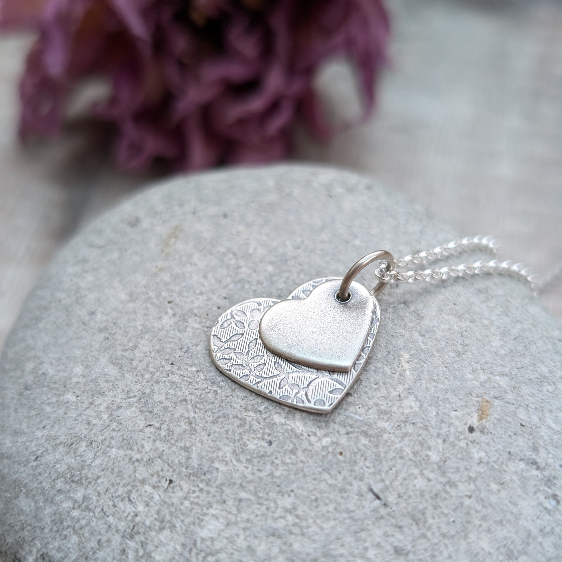 Pendant is a flat silver heart shape with detailed stamped floral pattern, overlapped by a smaller flat silver heart shape with softly brushed finish. The hearts have been oxidised to enhance the floral details. Suspended from a silver chain via a small circle link. 