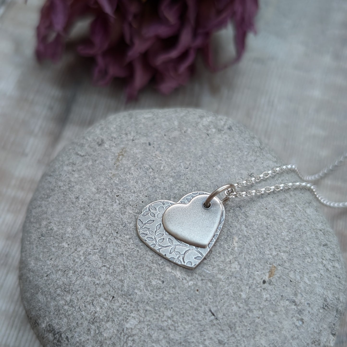 Pendant is a flat silver heart shape with detailed stamped floral pattern, overlapped by a smaller flat silver heart shape with softly brushed finish. The hearts have been oxidised to enhance the floral details. Suspended from a silver chain via a small circle link. 