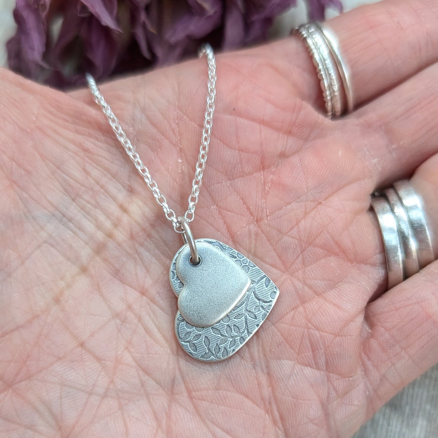 Pendant is a flat silver heart shape with detailed stamped floral pattern, overlapped by a smaller flat silver heart shape with softly brushed finish. The hearts have been oxidised to enhance the floral details. Suspended from a silver chain via a small circle link. 