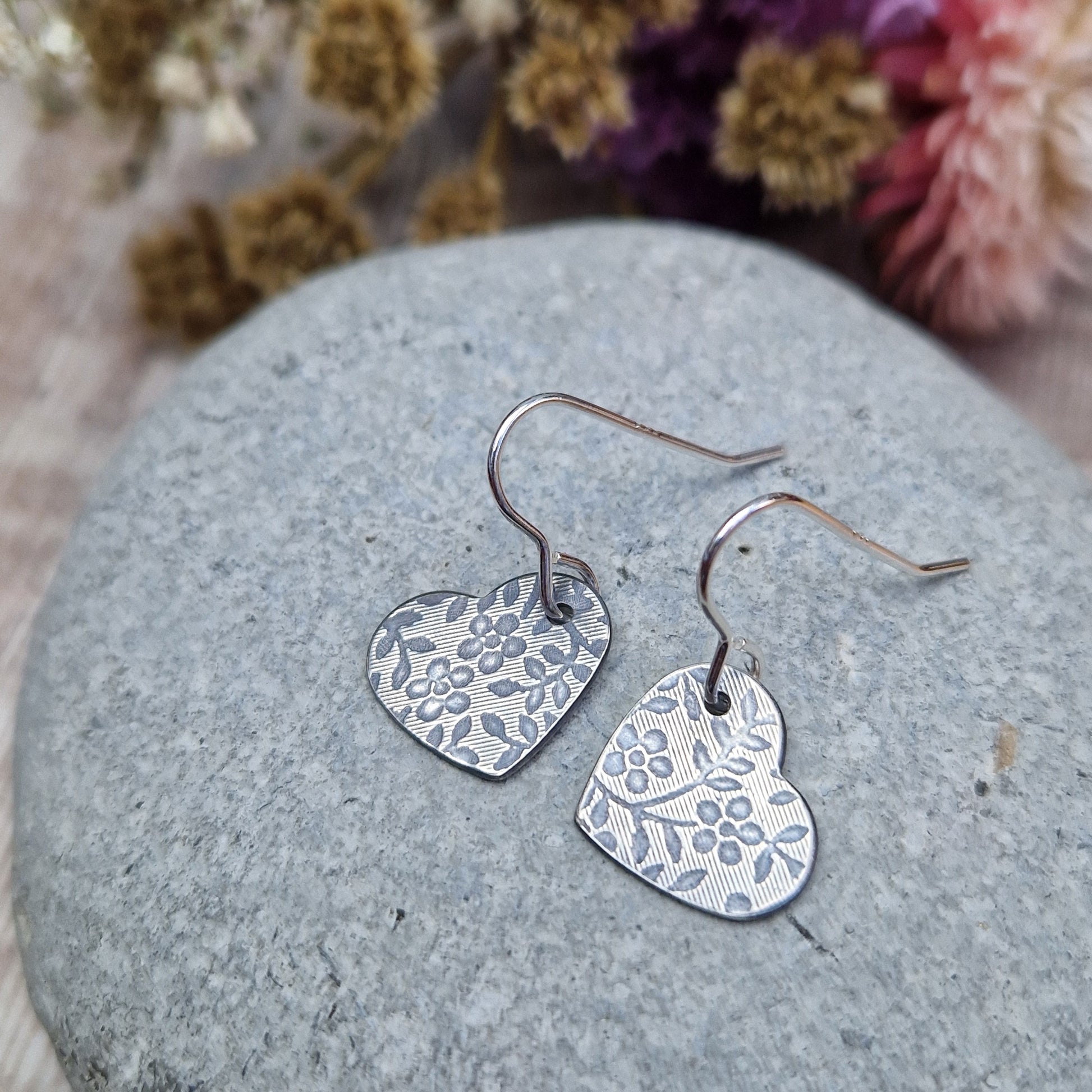 Each earring is an oxidised Sterling Silver heart shape hanging from silver earring wire. The heart shape is stamped with a floral pattern of flowers with 5 petals and leaves.