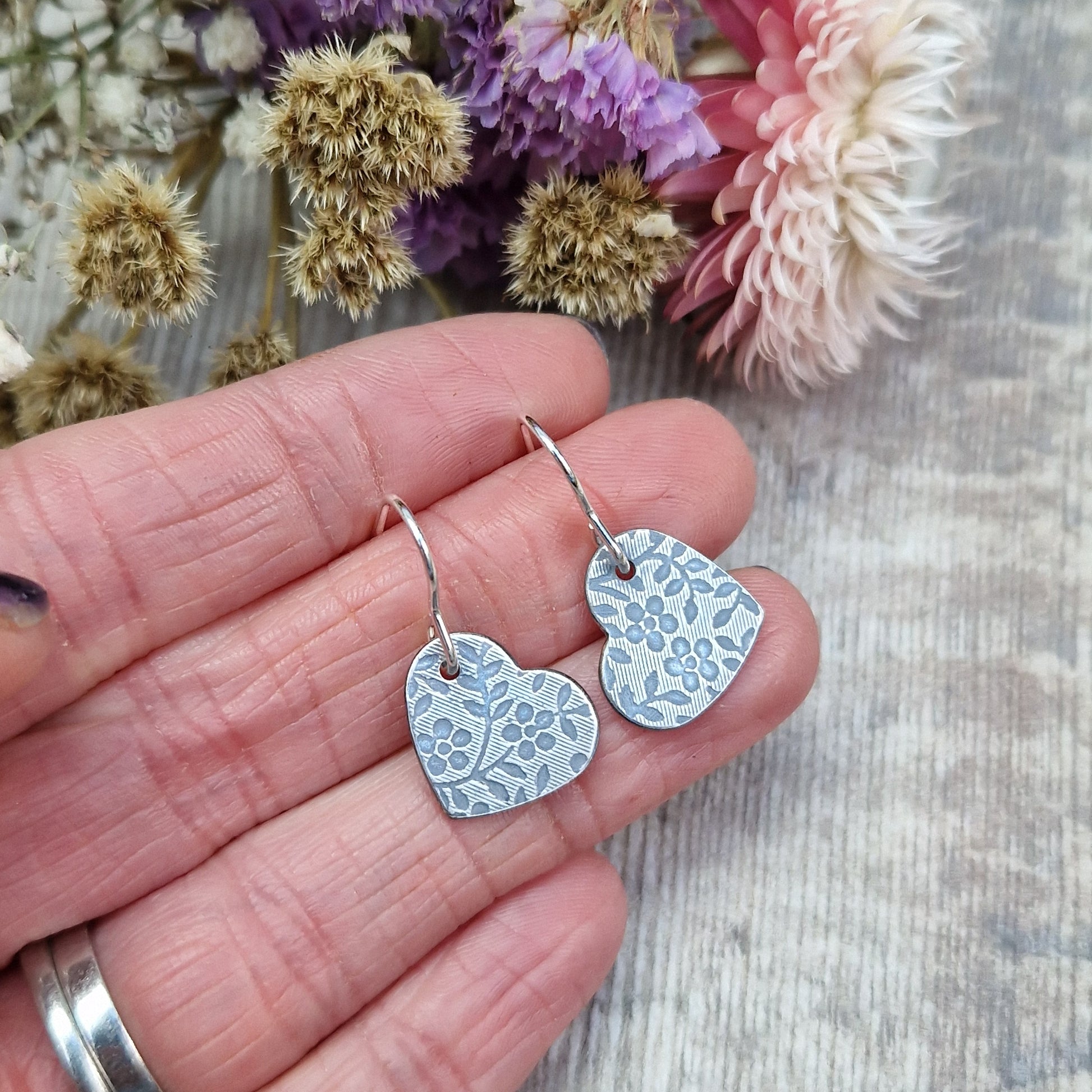 Each earring is an oxidised Sterling Silver heart shape hanging from silver earring wire. The heart shape is stamped with a floral pattern of flowers with 5 petals and leaves.