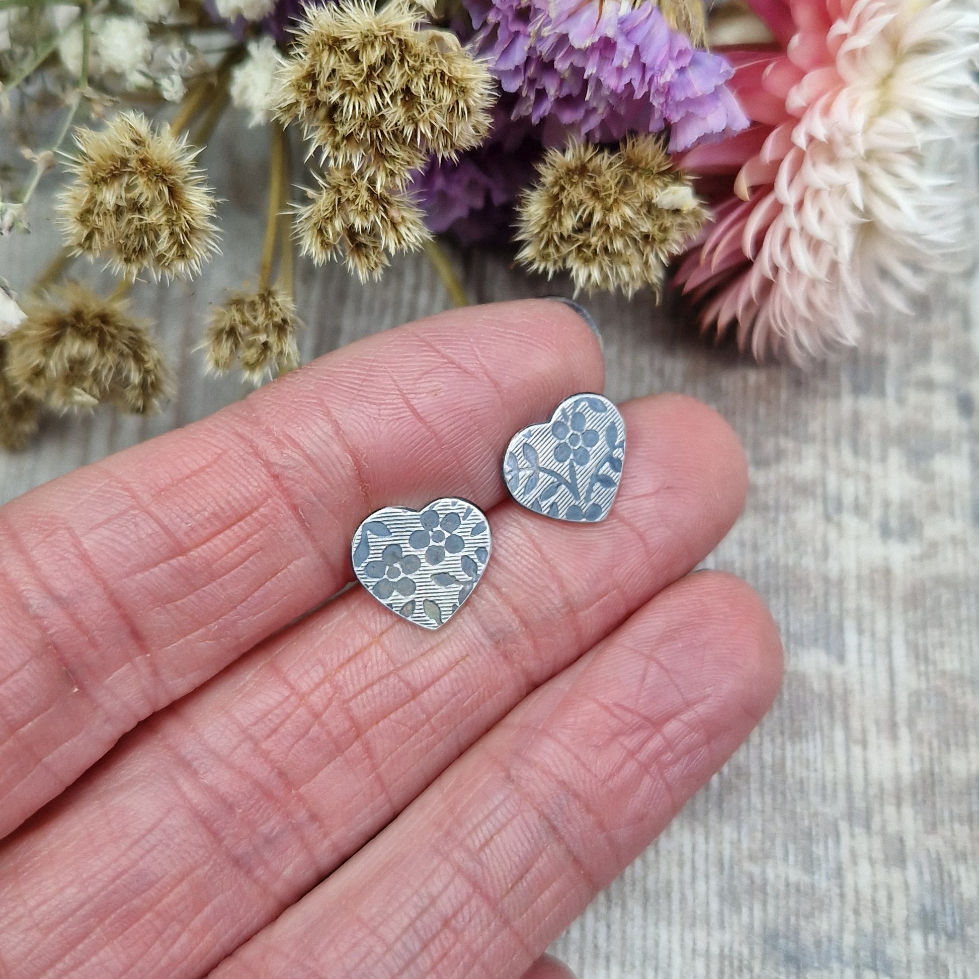 Each earring is an oxidised Sterling Silver heart shape on silver stud bar, stamped with a floral pattern of flowers with 5 petals and leaves. 