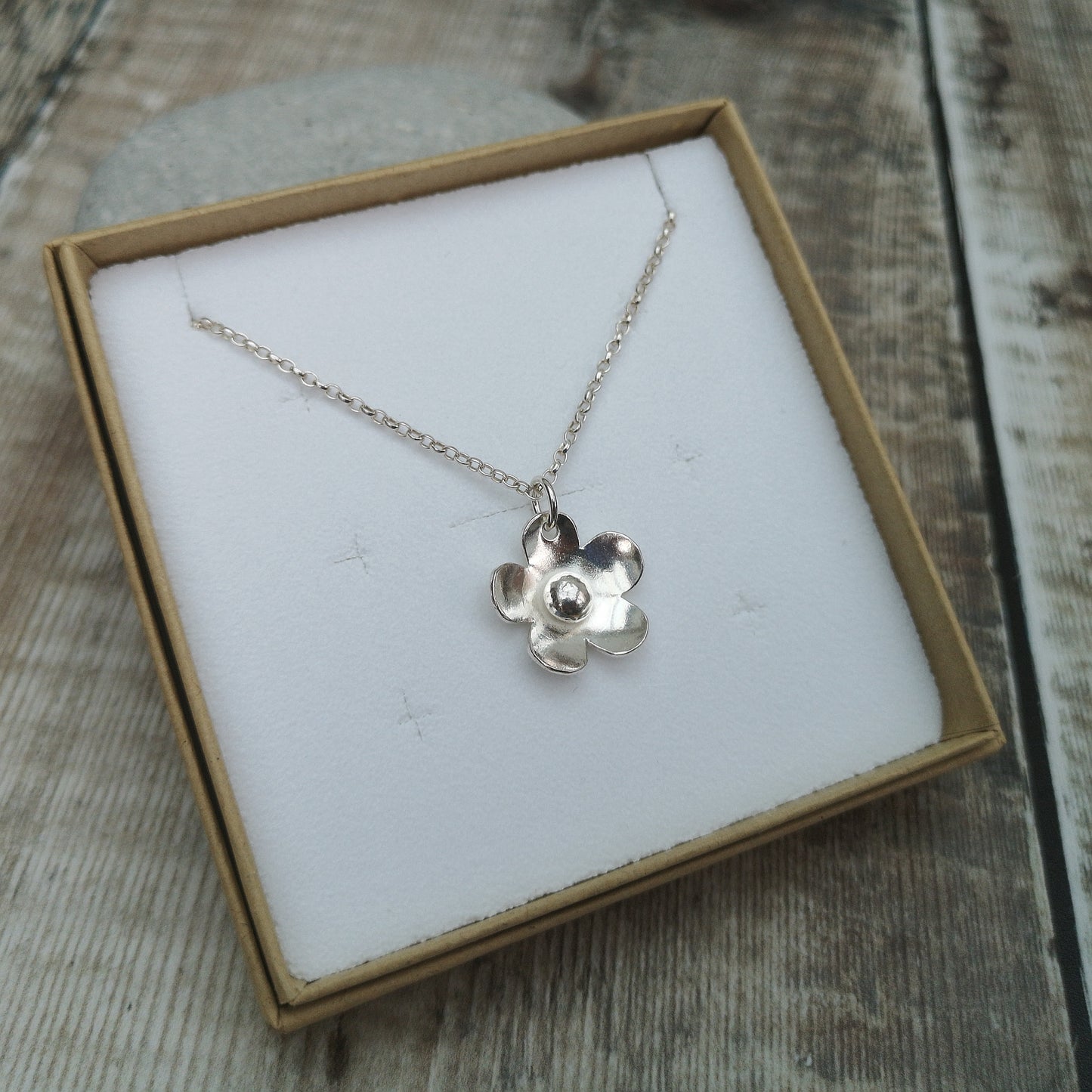 Sterling silver flower necklace displayed in a simple cardboard jewellery box with a white insert. The necklace features a polished flower pendant on a delicate silver chain, highlighting the handcrafted nature of the design.