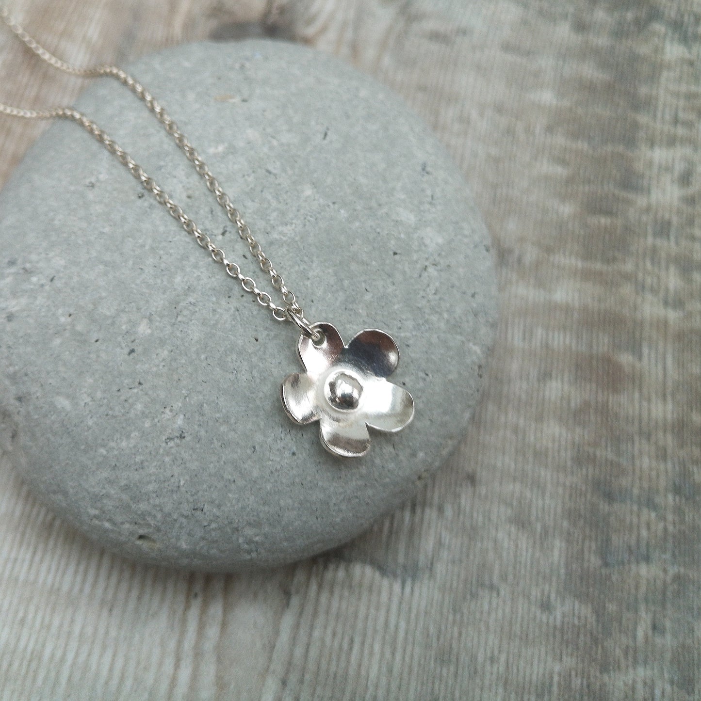 Close-up of a sterling silver flower necklace resting on a smooth grey stone. The necklace showcases a handcrafted flower pendant with softly rounded petals and a shiny, polished finish.