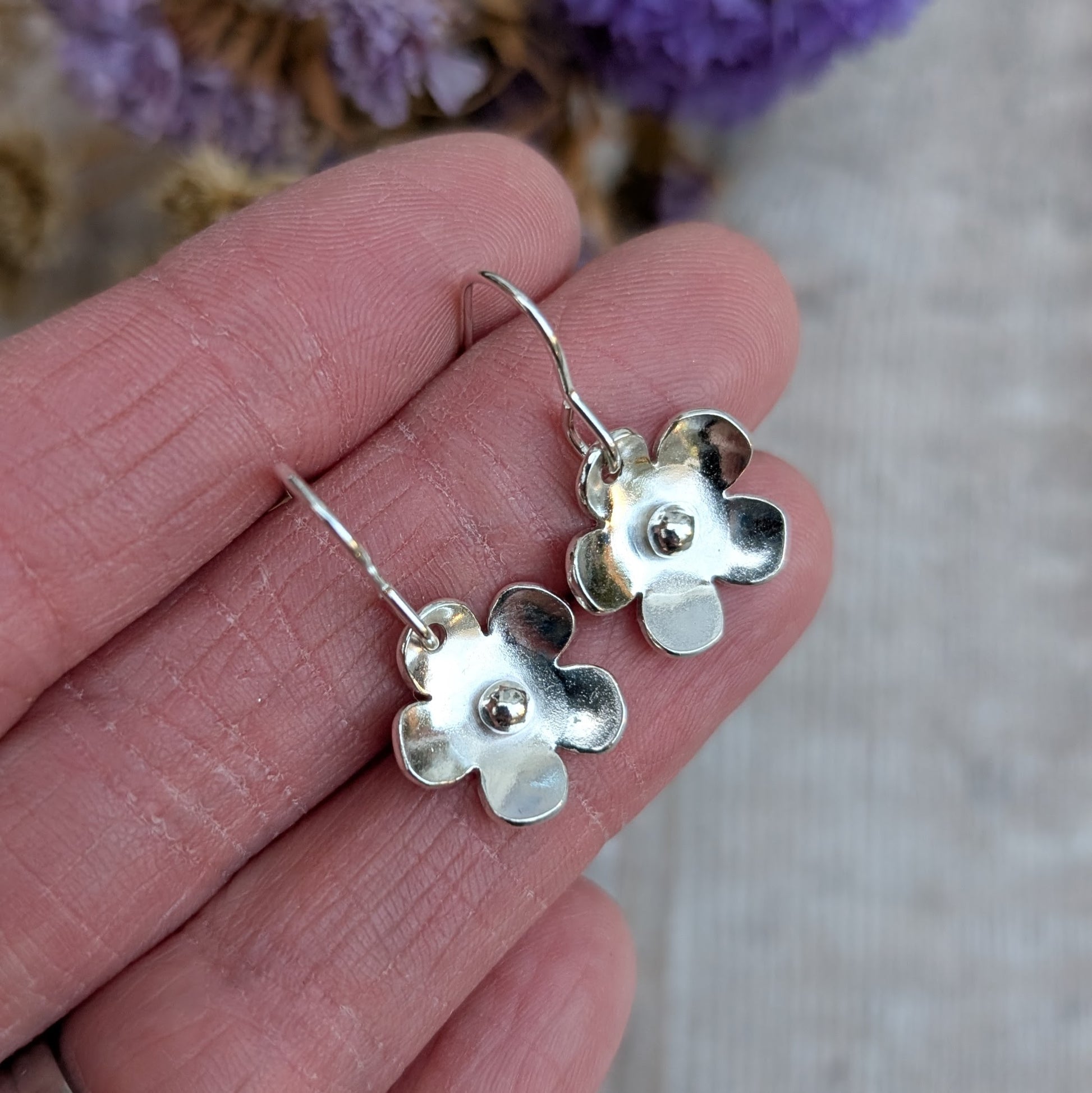 A pair of handmade sterling silver flower earrings with a small silver pebble at the center of each bloom, held between fingers. The earrings dangle from simple ear wires, and a background of dried flowers adds a soft, natural touch.