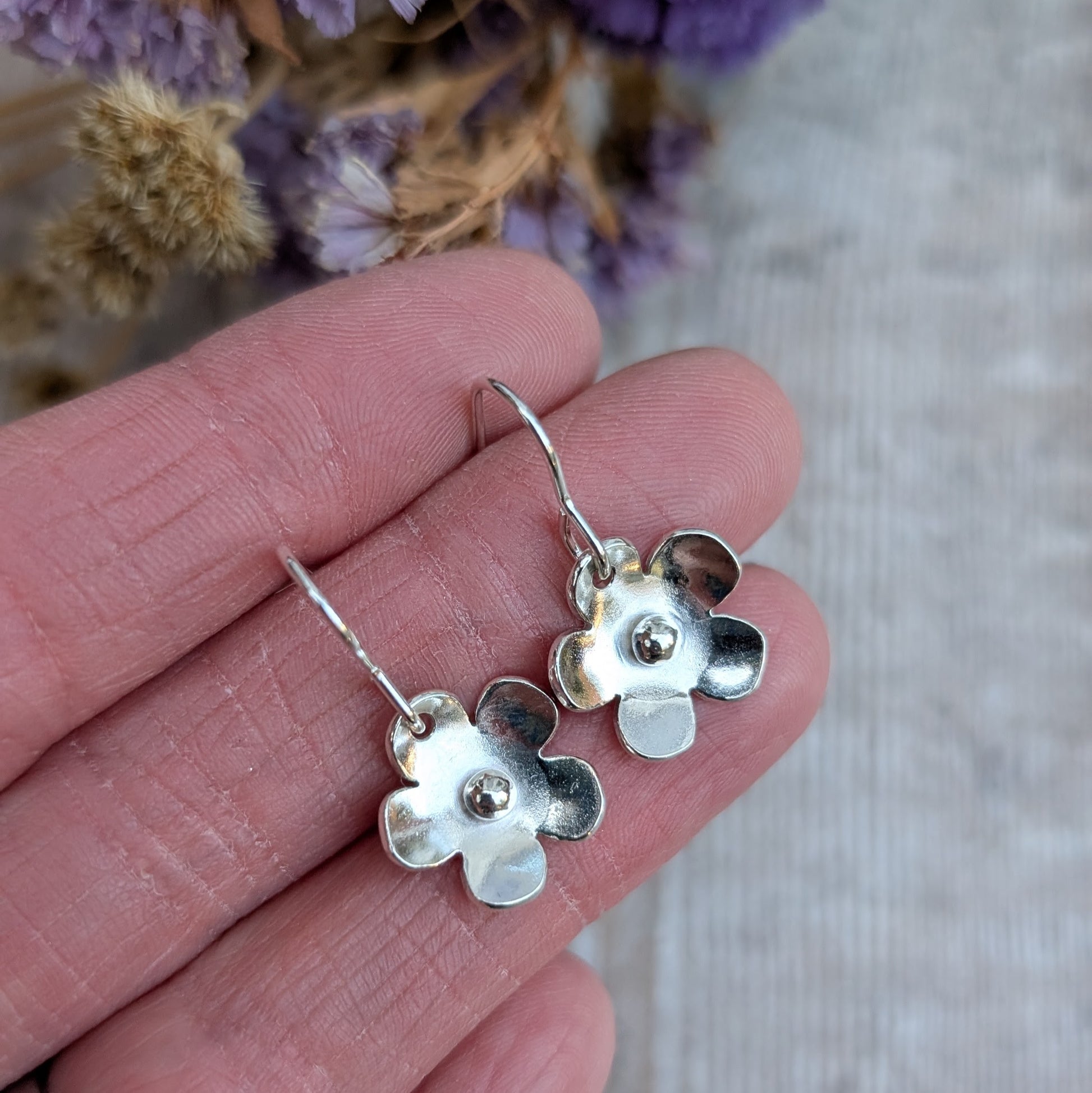 A pair of handmade sterling silver flower earrings with a small silver pebble at the center of each bloom, held between fingers. The earrings dangle from simple ear wires, and a background of dried flowers adds a soft, natural touch.