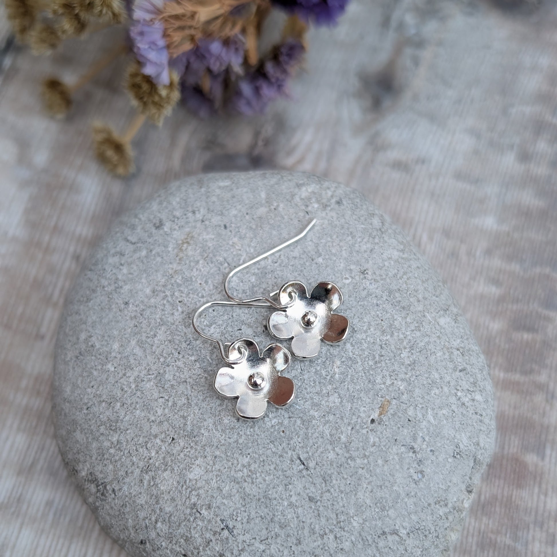 The silver flower earrings resting on a smooth grey stone. The slightly curved petals catch the light, and the silver pebbles at their centers add a subtle texture. Dried purple flowers in the background complement the scene.