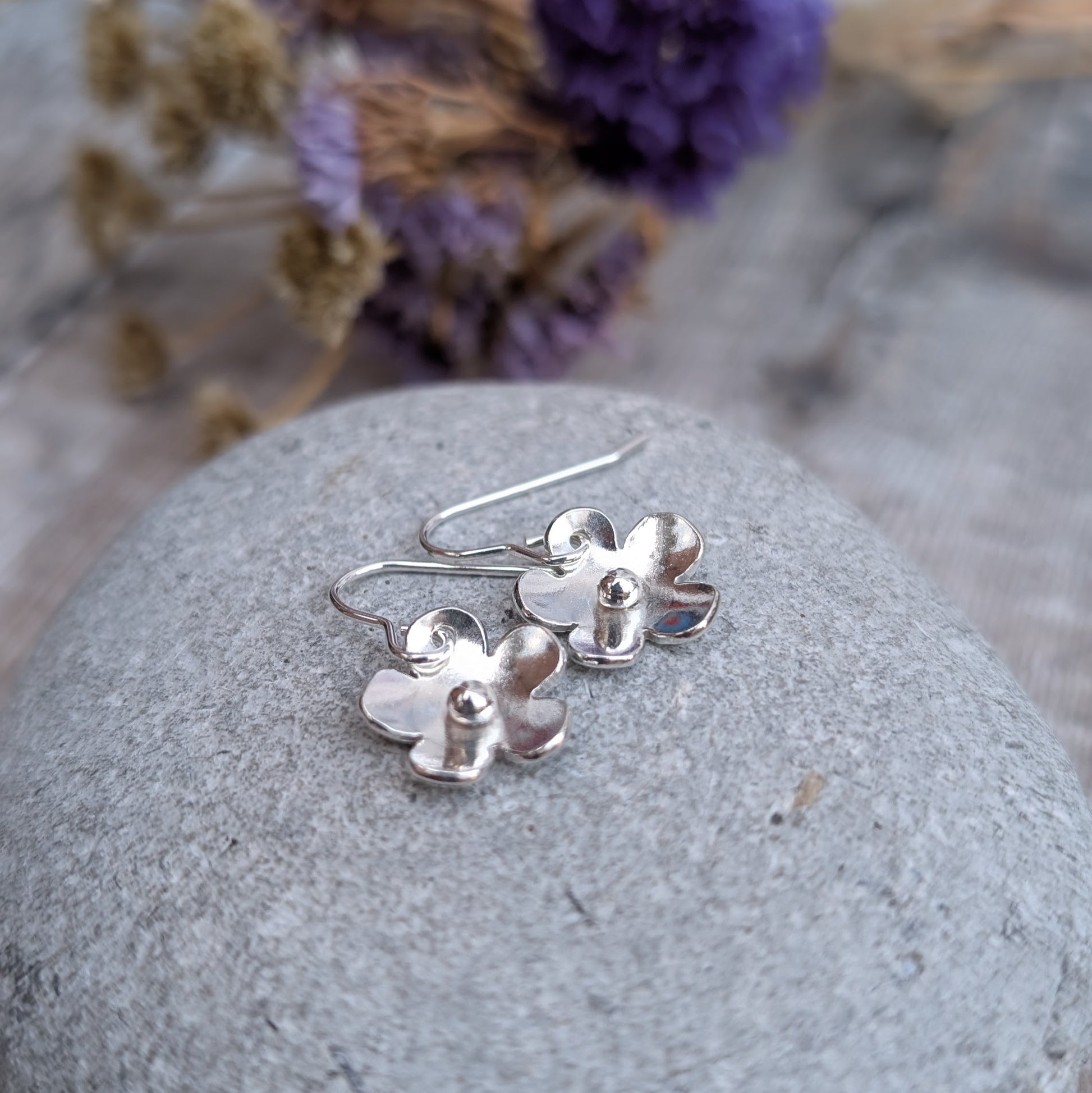 The silver flower earrings resting on a smooth grey stone. The slightly curved petals catch the light, and the silver pebbles at their centers add a subtle texture. Dried purple flowers in the background complement the scene.