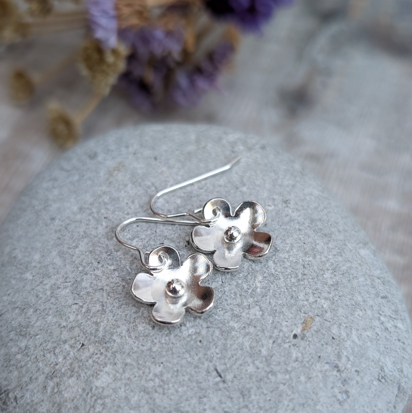 The silver flower earrings resting on a smooth grey stone. The slightly curved petals catch the light, and the silver pebbles at their centers add a subtle texture. Dried purple flowers in the background complement the scene.