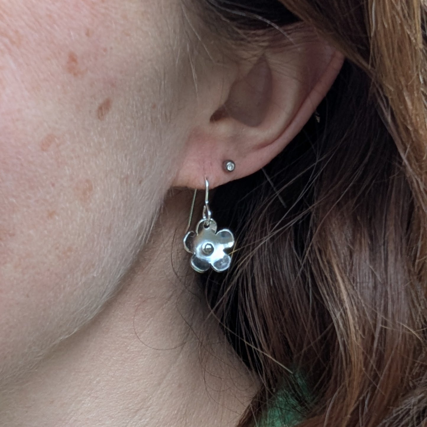 A close-up of one silver flower earring being worn. The earring dangles just below the earlobe, with its gently domed petals reflecting light. A small silver stud is visible in the second piercing, and soft brown hair frames the ear.