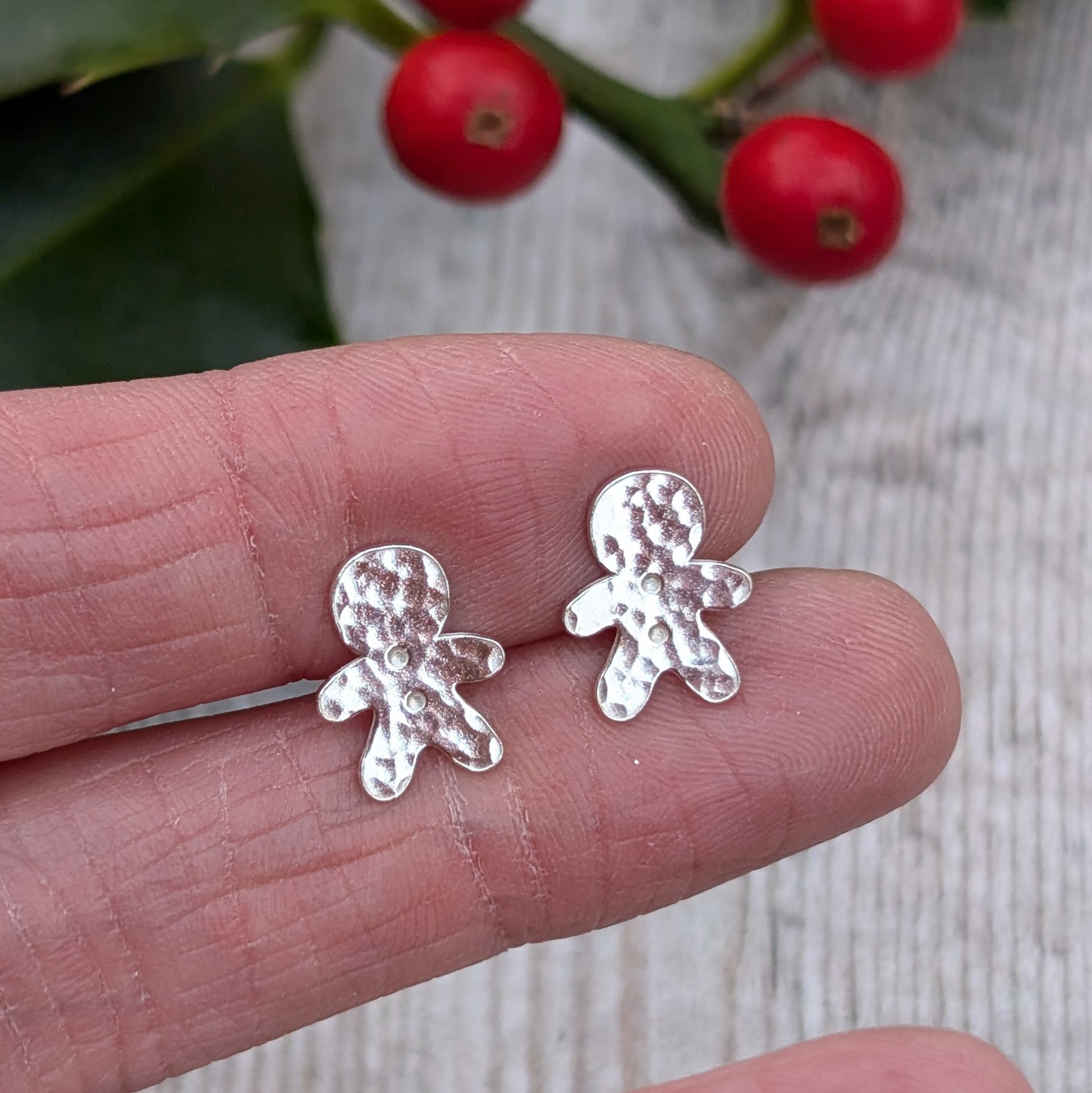 Close-up of a hand holding a pair of small silver gingerbread man stud earrings with a hammered texture. Bright red holly berries are visible in the background.