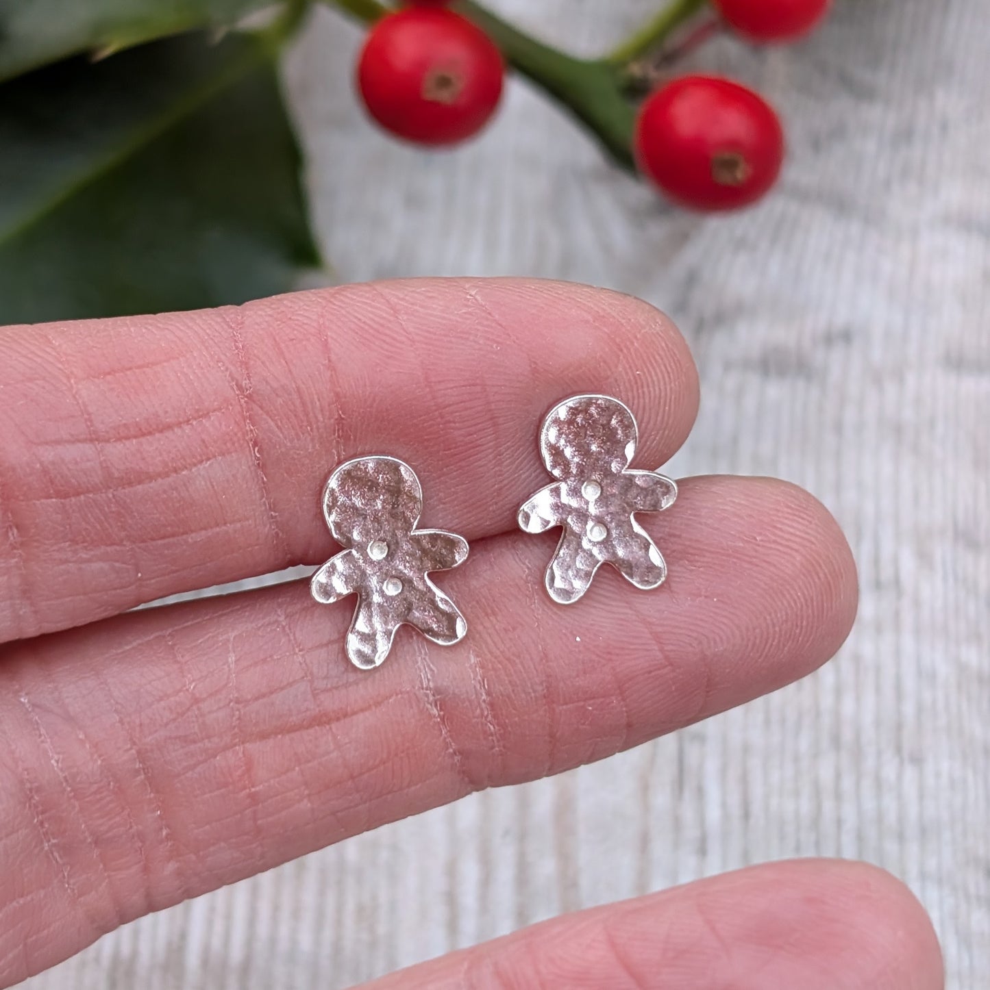 Close-up of a hand holding a pair of small silver gingerbread man stud earrings with a hammered texture. Bright red holly berries are visible in the background.