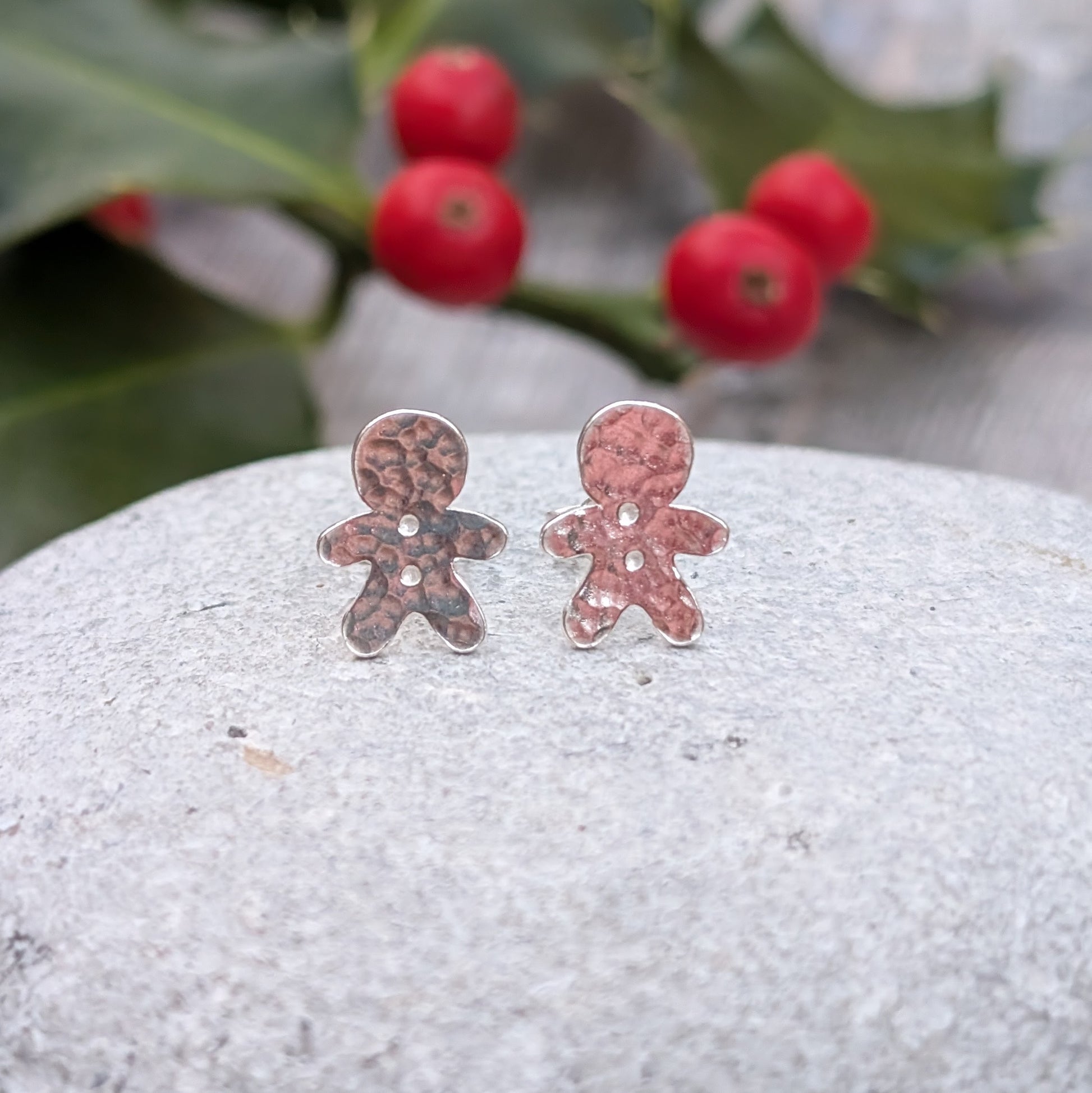 Two small silver gingerbread man stud earrings with a hammered texture placed on a grey stone. Red holly berries and green leaves are softly blurred in the background.