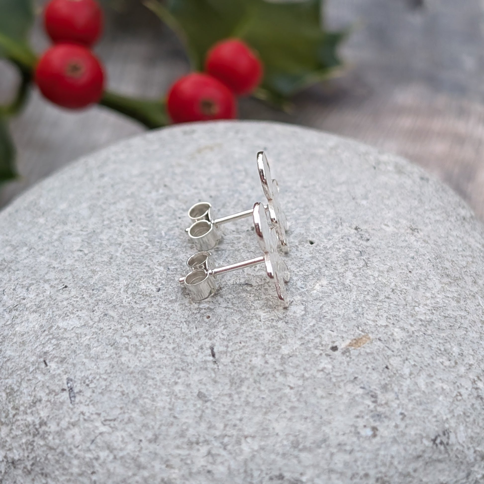 Side view of two silver gingerbread man stud earrings lying flat on a grey stone, showing the earring posts and butterfly backings. Red holly berries are blurred in the background.