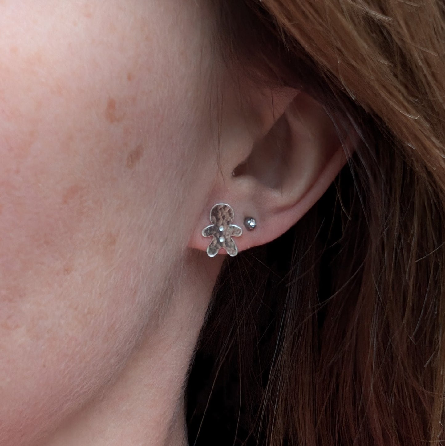 A silver gingerbread man stud earring worn on an earlobe, showing its small size and hammered texture. Another small silver stud earring is worn above it.