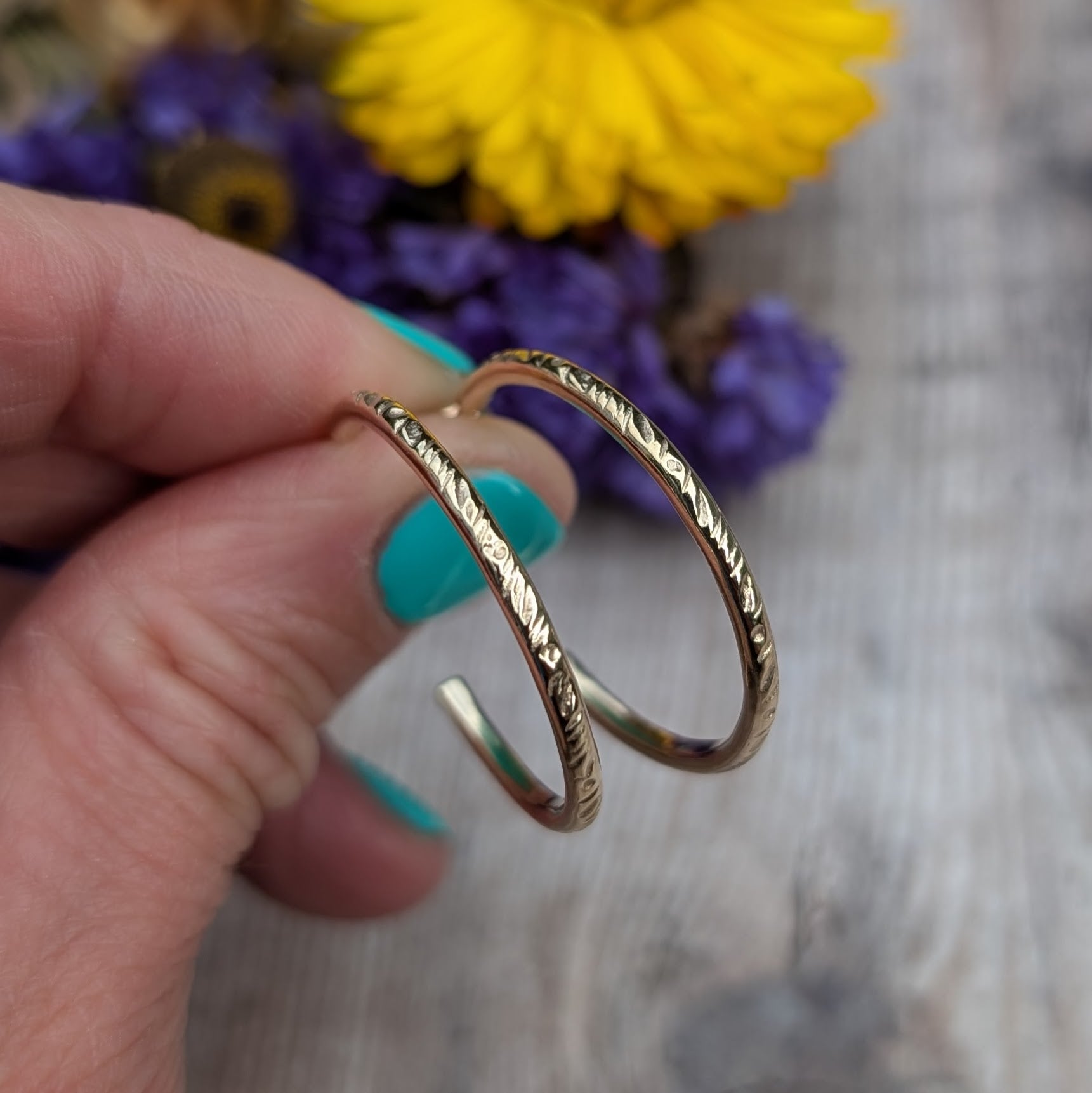 Close-up view of handmade gold hoop earrings with a delicate 'Meadow' pattern, crafted from recycled 9ct gold wire, held against a wooden background with flowers in the distance