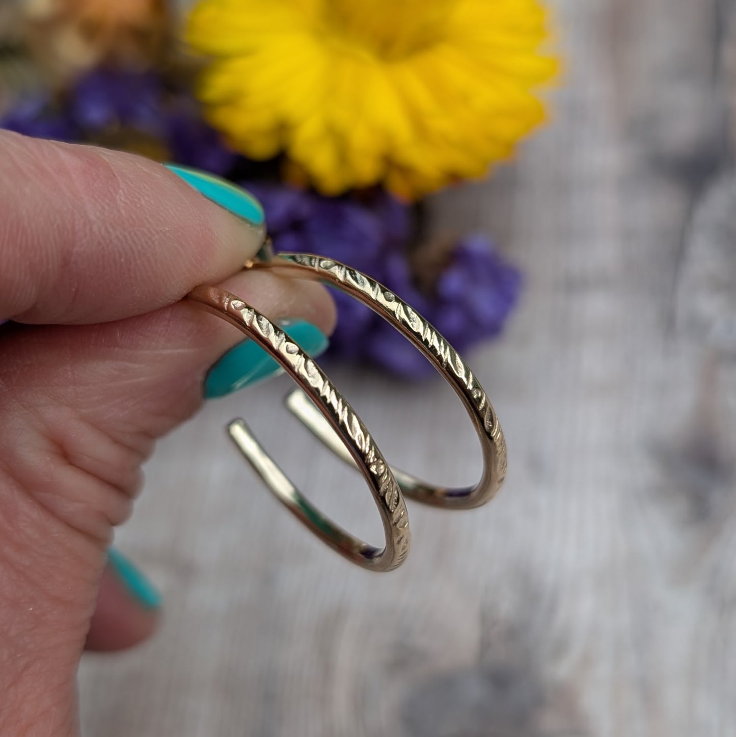 Close-up view of handmade gold hoop earrings with a delicate 'Meadow' pattern, crafted from recycled 9ct gold wire, held against a wooden background with flowers in the distance.