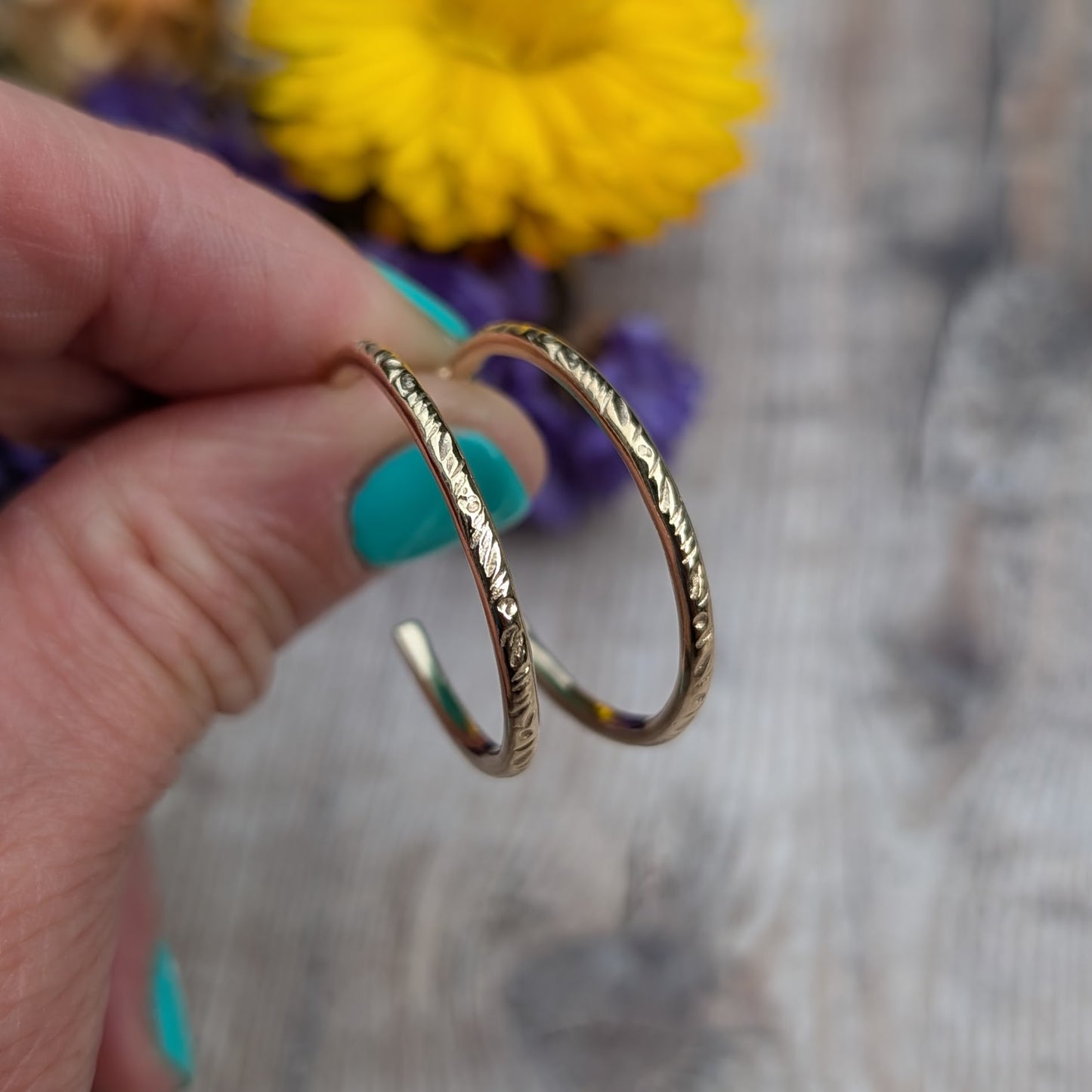 Close-up view of handmade gold hoop earrings with a delicate 'Meadow' pattern, crafted from recycled 9ct gold wire, held against a wooden background with flowers in the distance