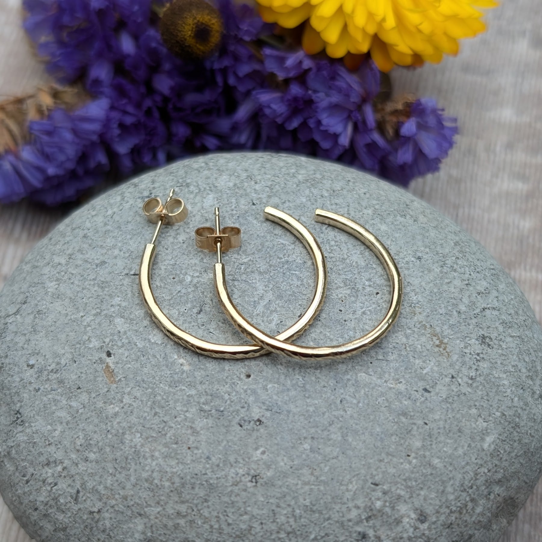 Pair of gold hoop earrings featuring the 'Meadow' pattern, displayed on a smooth stone with purple and yellow flowers in the background, highlighting the handcrafted details and recycled 9ct gold material.