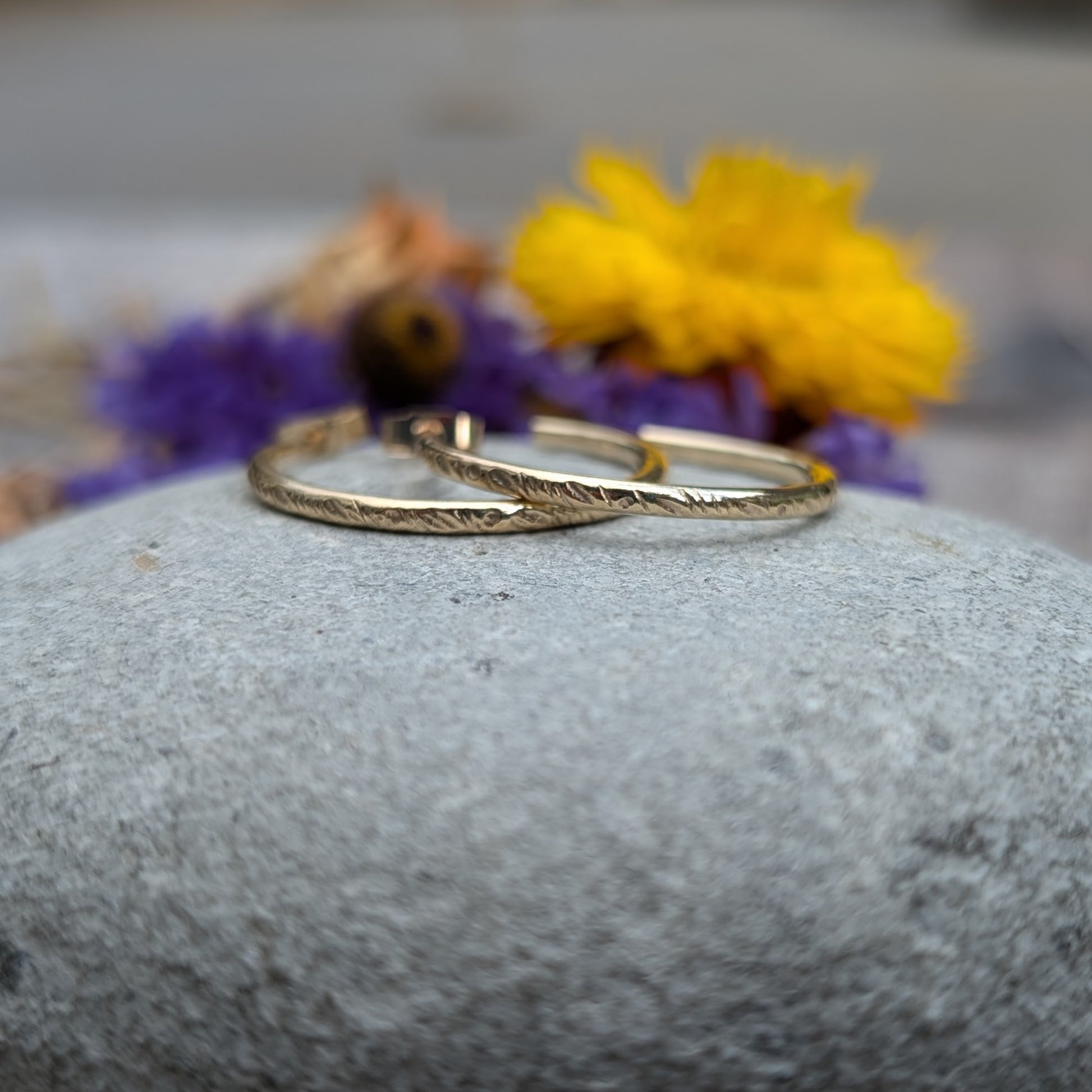 Pair of gold hoop earrings featuring the 'Meadow' pattern, displayed on a smooth stone with purple and yellow flowers in the background, highlighting the handcrafted details and recycled 9ct gold material.