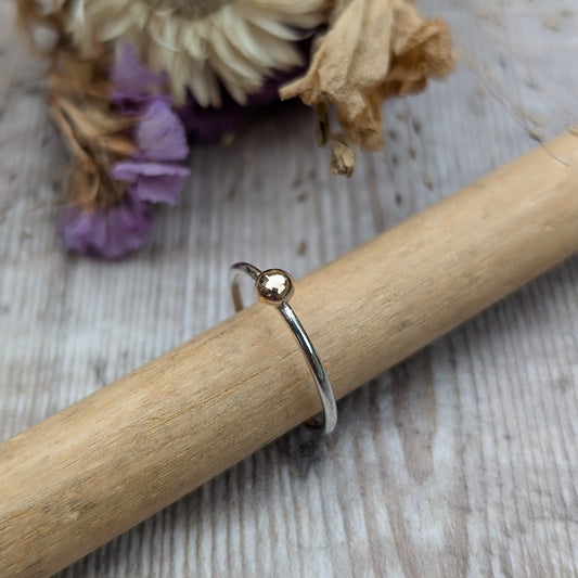 A close-up of a delicate ring on a wooden dowel. The ring features a recycled silver band with a small recycled 9ct gold pebble as its focal point, highlighting its minimalistic and elegant design.