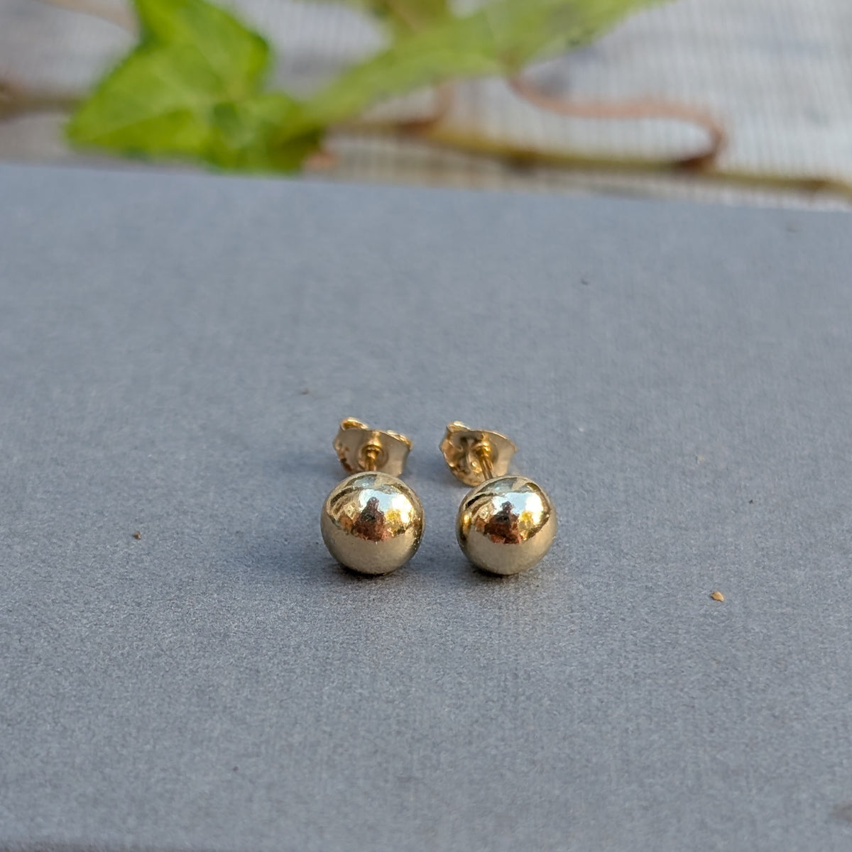 Pair of handmade solid gold pebble stud earrings sitting on a grey background with green ivy behind.
