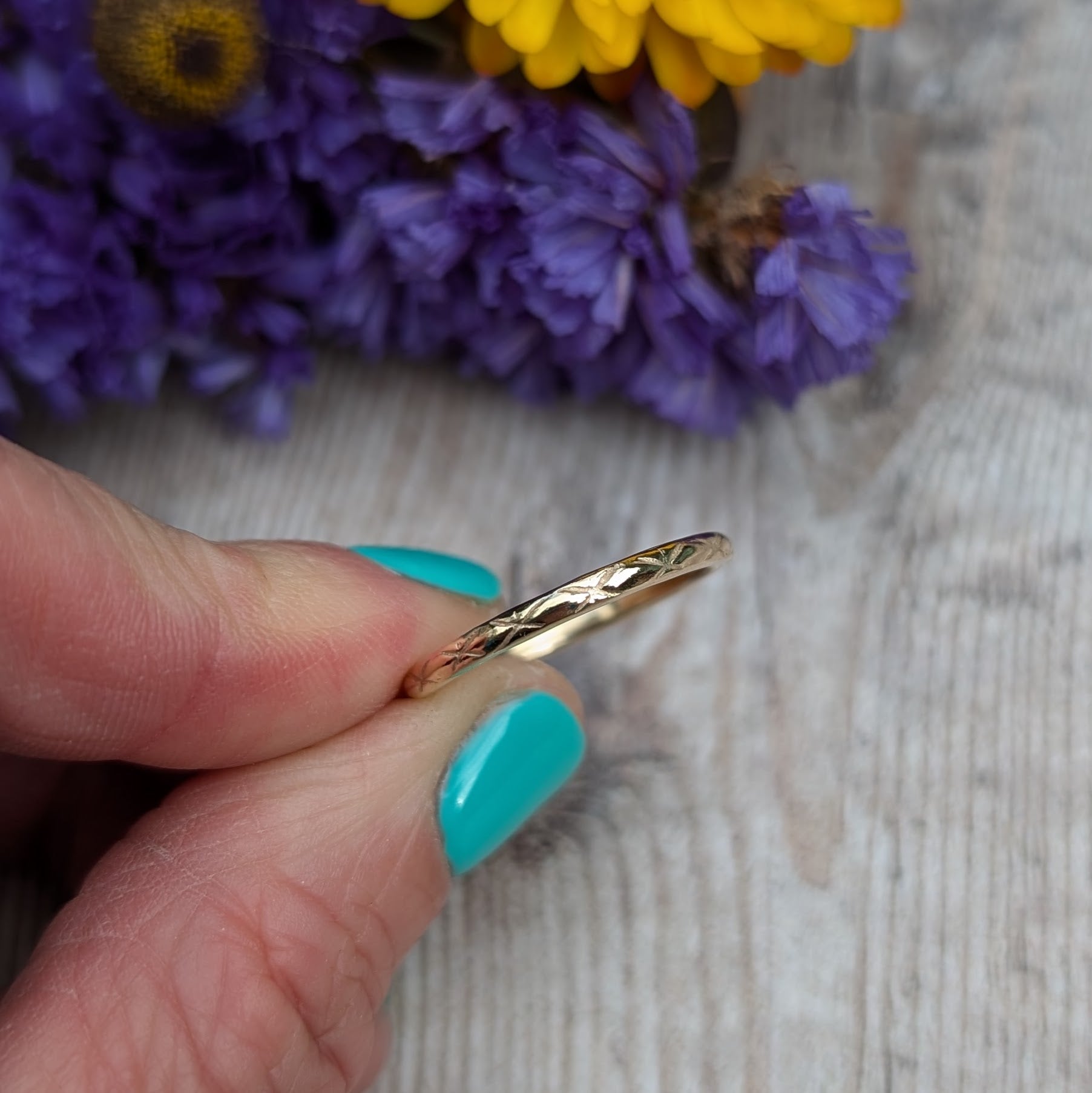 A hand holding a thin gold ring against a wooden surface with purple and yellow flowers in the background. The ring showcases a 'Starlight' pattern, carefully crafted by hand, with a polished finish that highlights the intricate design.