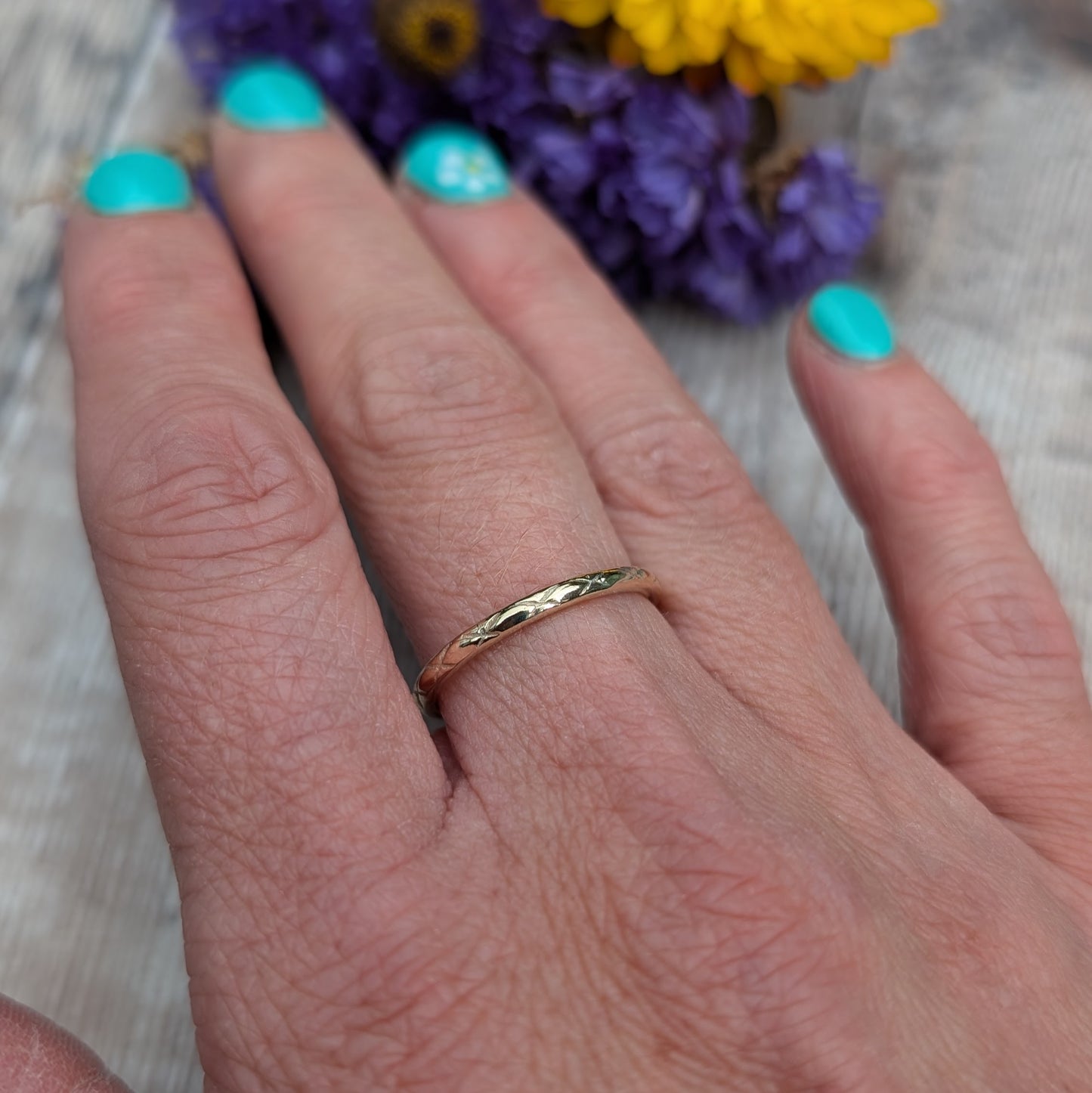 A close-up image of a hand wearing a thin gold ring with a delicate pattern. The ring features the 'Starlight' pattern, created by impressing the design by hand. The background includes purple and yellow flowers, adding a vibrant touch to the image.