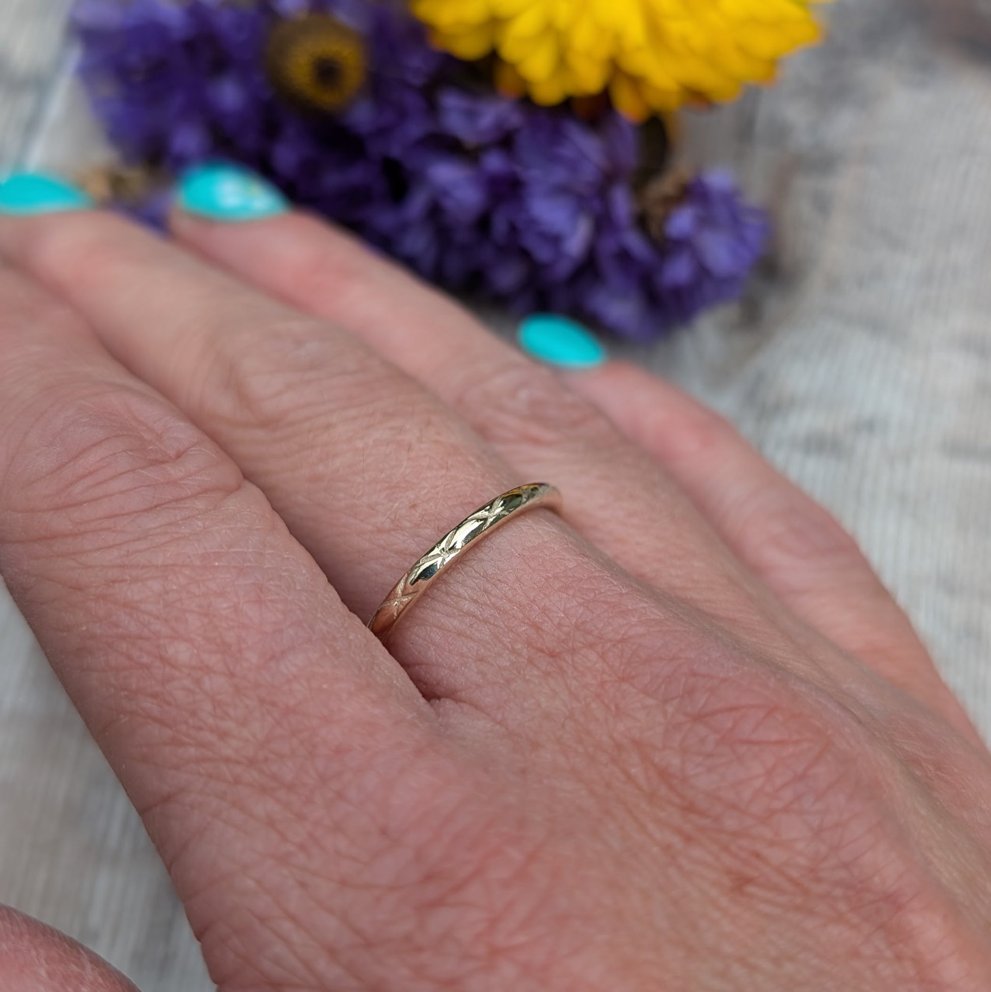 A close-up image of a hand wearing a thin gold ring with a delicate pattern. The ring features the 'Starlight' pattern, created by impressing the design by hand. The background includes purple and yellow flowers, adding a vibrant touch to the image.