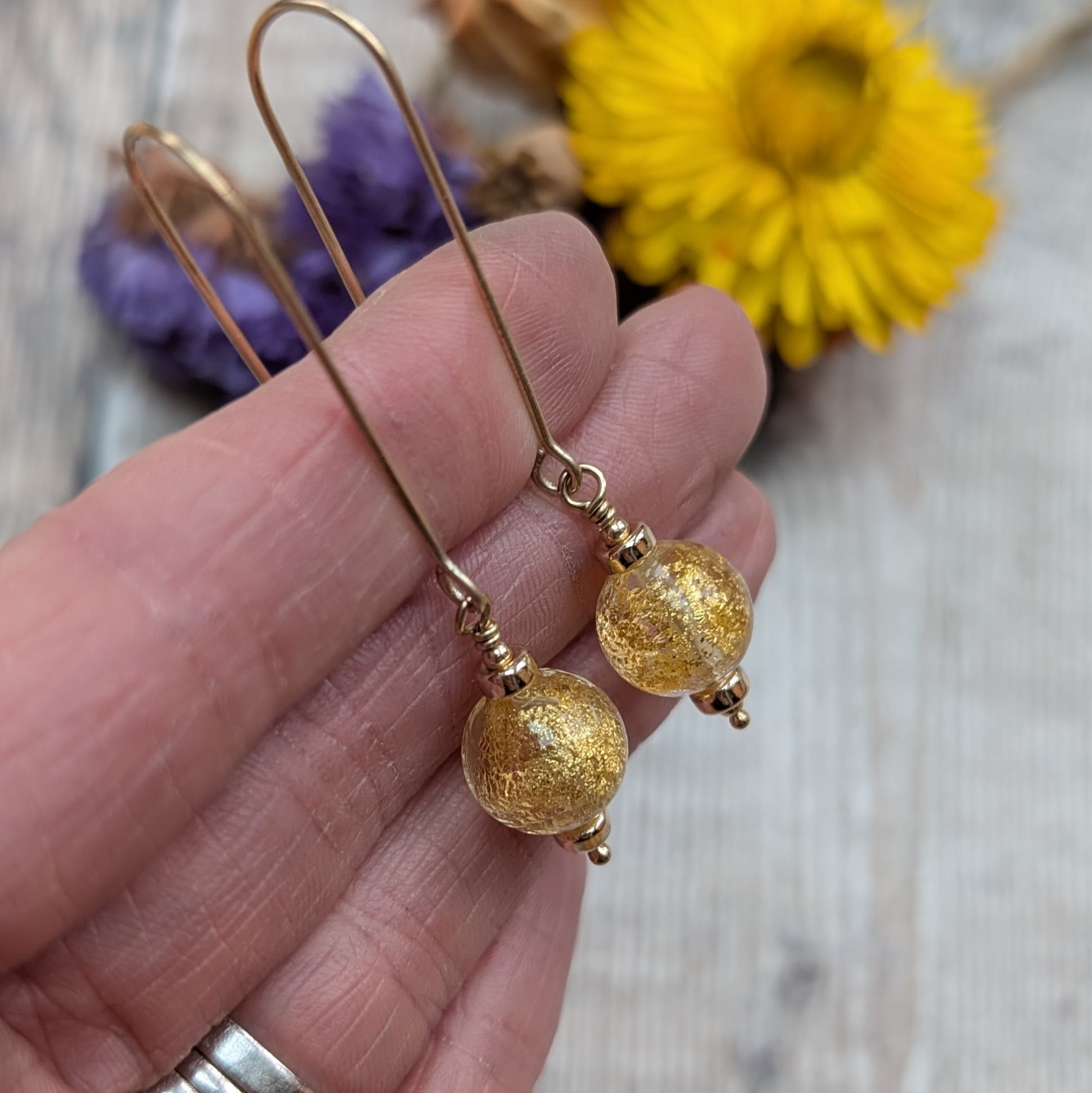 Detailed view of gold filled long hook earrings with spherical gold fleck Venetian glass beads, photographed against a light wooden surface with dried flowers.