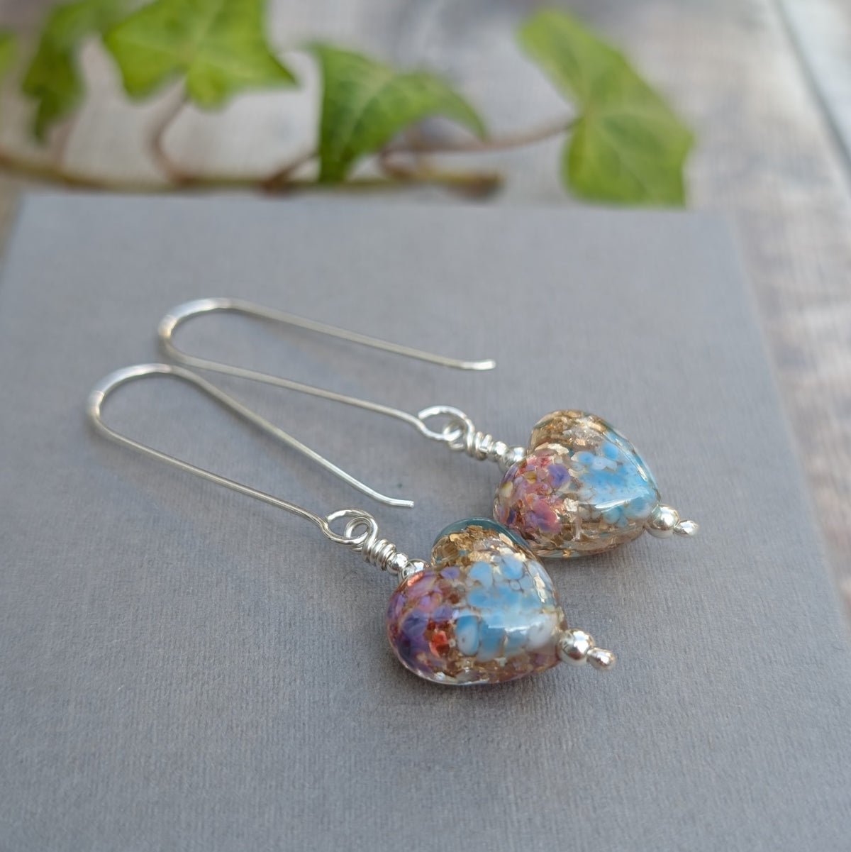 golden fantasy lampwork glass heart long silver earrings lying on a grey background with green ivy behind.