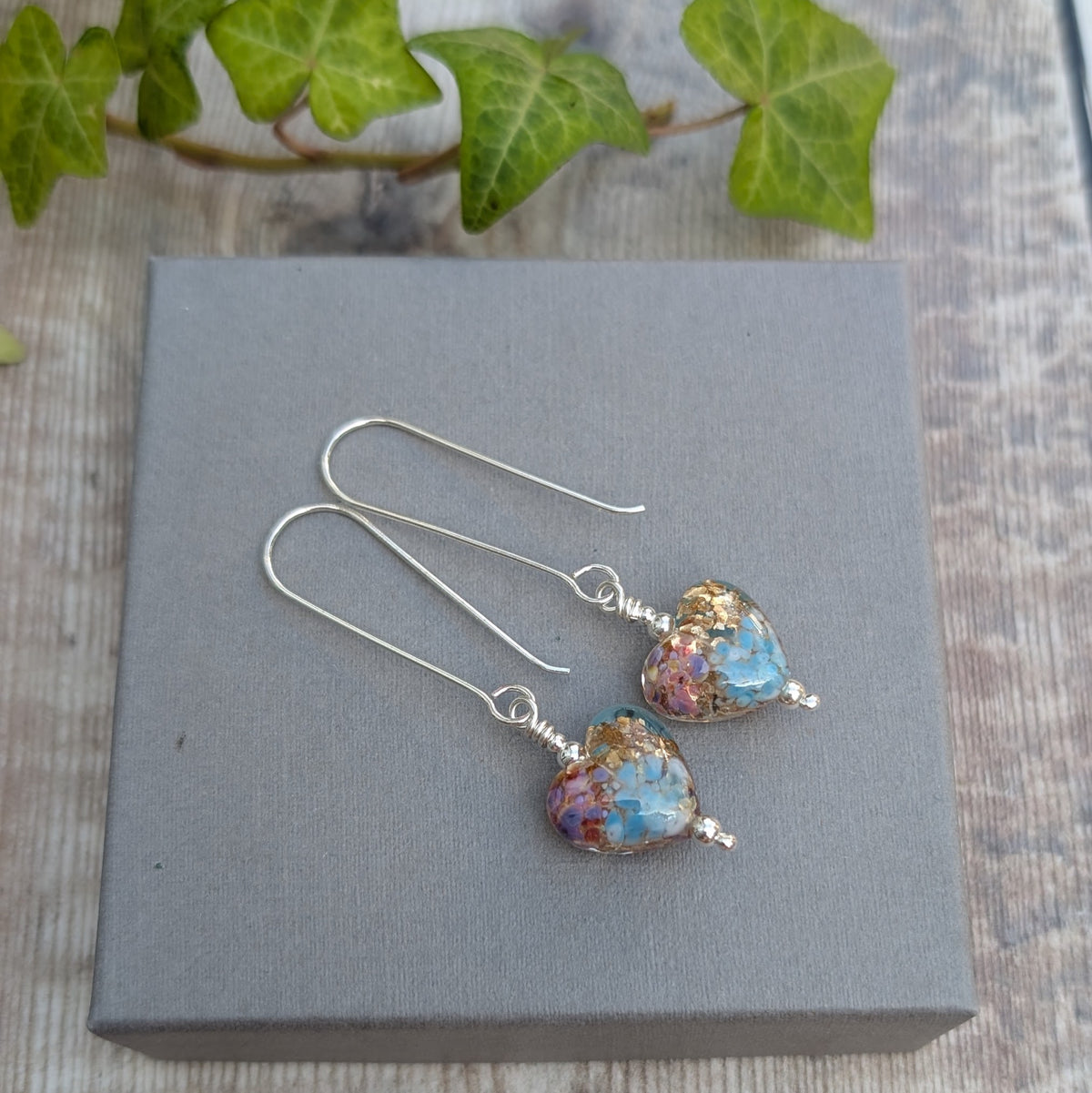 golden fantasy lampwork glass heart long silver earrings displayed on a grey jewellery box with green ivy in the background.