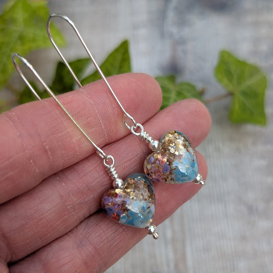 golden fantasy lampwork glass heart long silver earrings held in a hand showing the blue, pink, purple and gold glass heart beads.