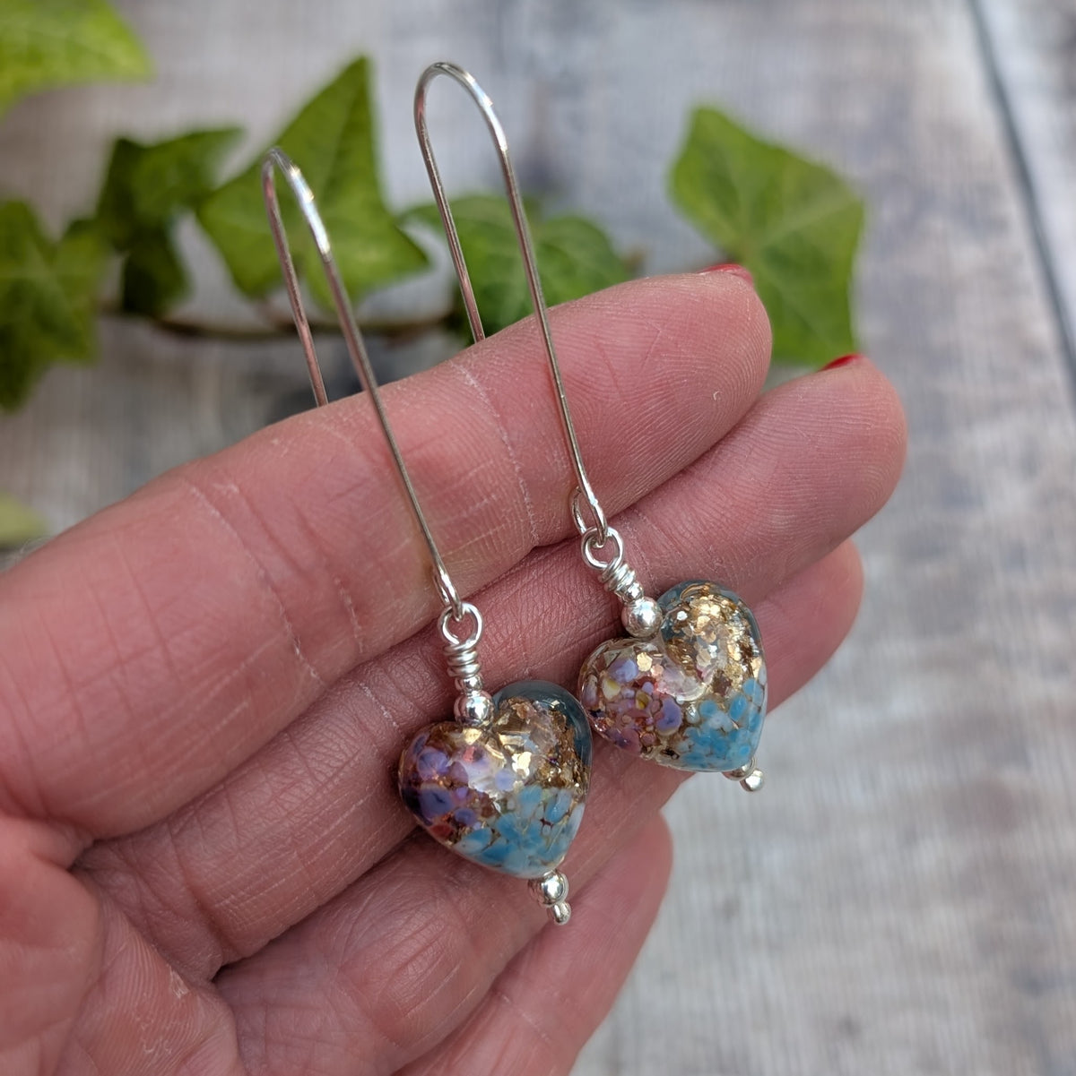 Close up of golden fantasy lampwork glass heart long silver earrings held in a hand showing the handmade silver ear wires and colourful glass hearts.