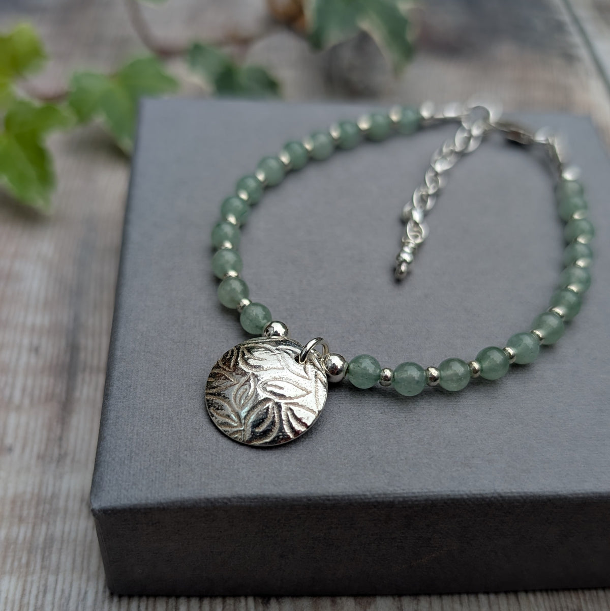 Green aventurine and silver leaf disc bracelet handmade with recycled sterling silver, featuring pale green gemstone beads, silver spacers and a textured leaf charm, displayed on a grey box.