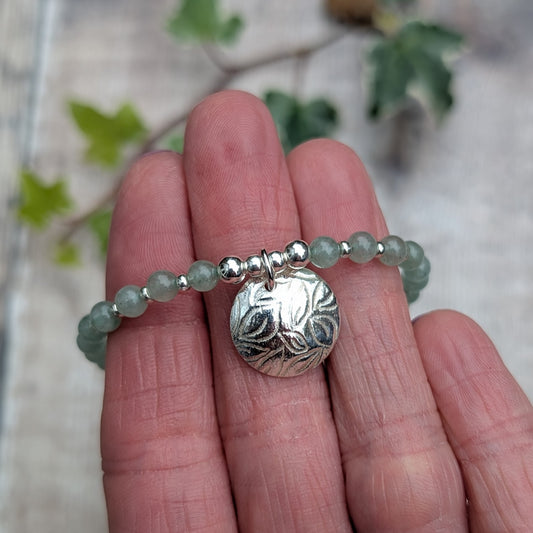 Close up of green aventurine and silver leaf disc bracelet showing the handmade sterling silver leaf patterned charm and green gemstone beads held against fingers.