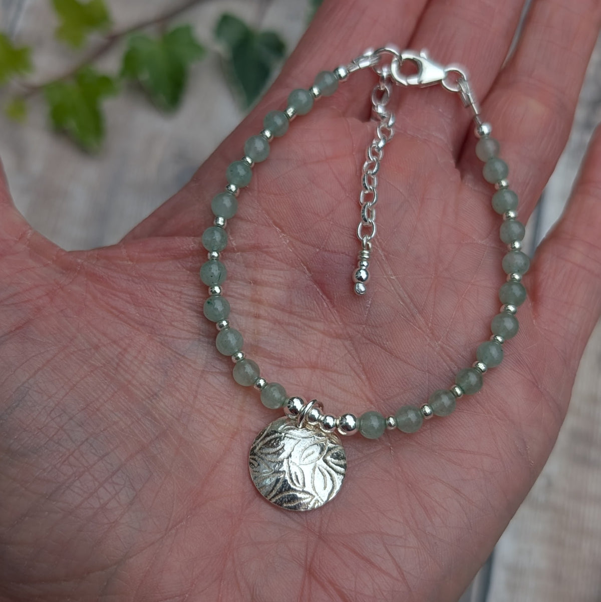 Green aventurine and silver leaf disc bracelet displayed in the palm of a hand, showing the full beaded design, handmade silver leaf charm, lobster clasp and adjustable extender chain.