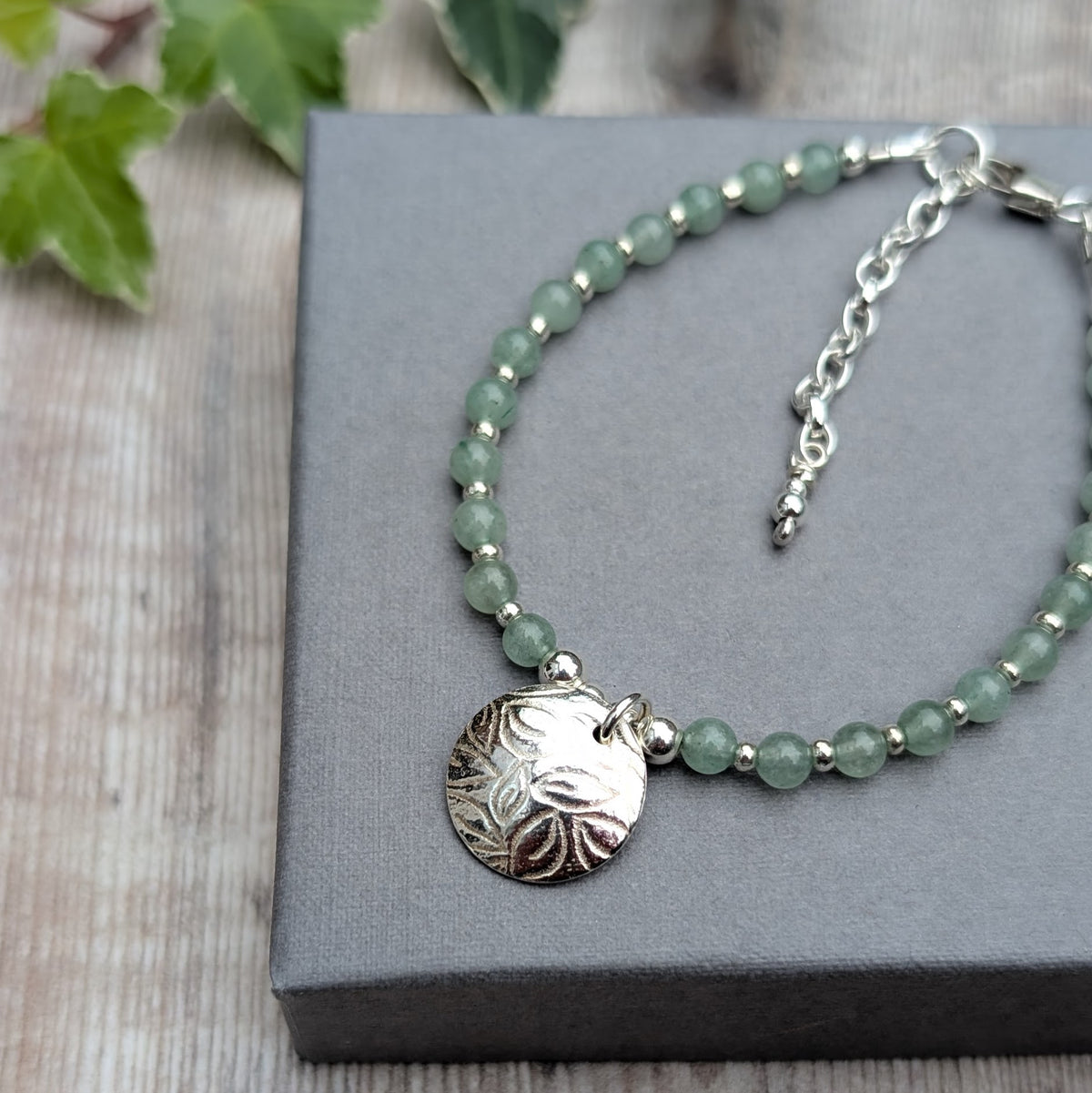 Green aventurine and silver leaf disc bracelet resting on a grey jewellery box, featuring pale green aventurine beads, sterling silver spacers and a round textured leaf charm.