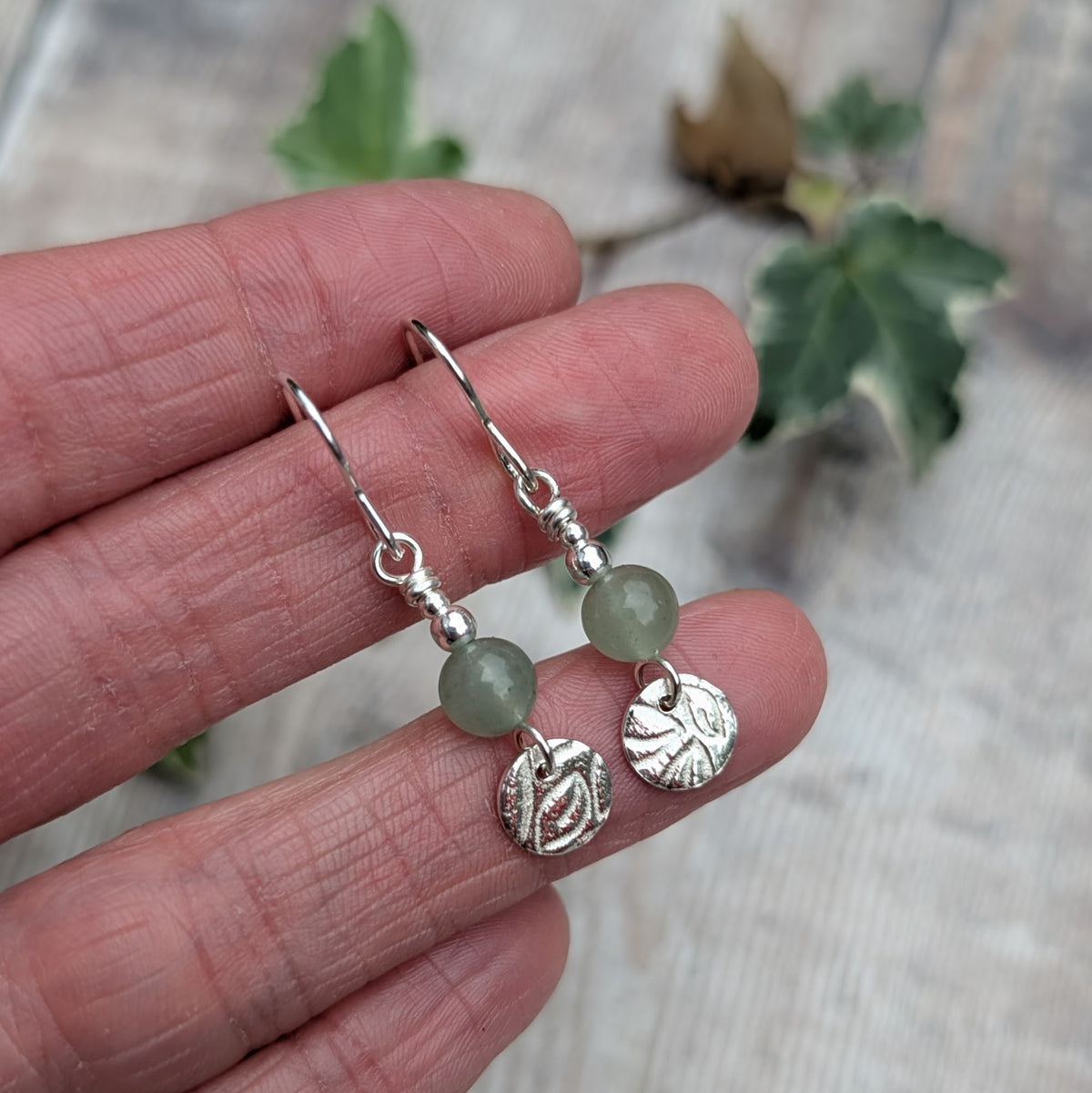 Close up of green aventurine and silver leaf earrings held in a hand, showing the detailed leaf pattern on the recycled sterling silver discs and soft green aventurine beads.