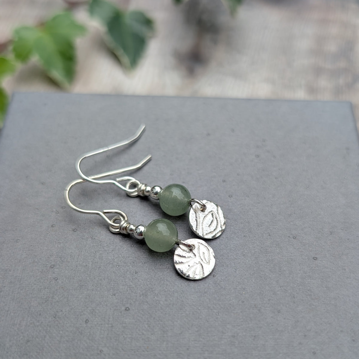 Green aventurine and silver leaf earrings laid flat on a grey background, showing handmade sterling silver ear wires, pale green gemstone beads and textured silver discs.