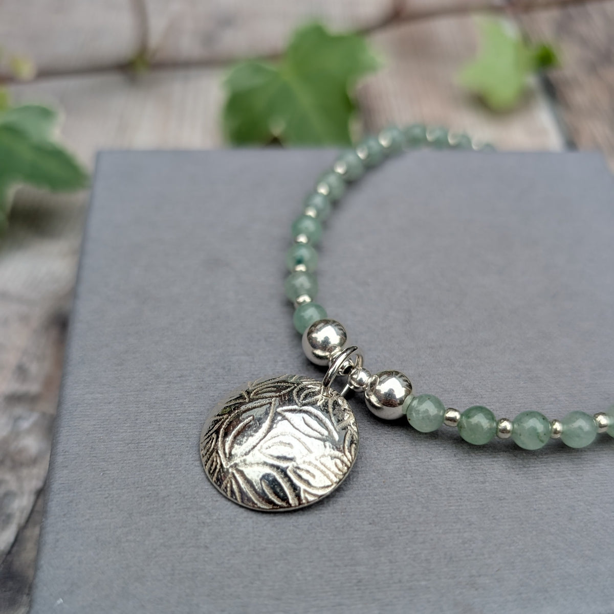 Close up of Green Aventurine and Silver Leaf Disc Necklace showing the textured sterling silver leaf charm, polished silver beads and pale green aventurine gemstones on a grey background.
