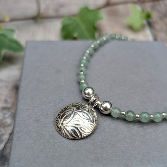Close up of Green Aventurine and Silver Leaf Disc Necklace showing the textured sterling silver leaf charm, polished silver beads and pale green aventurine gemstones on a grey background.