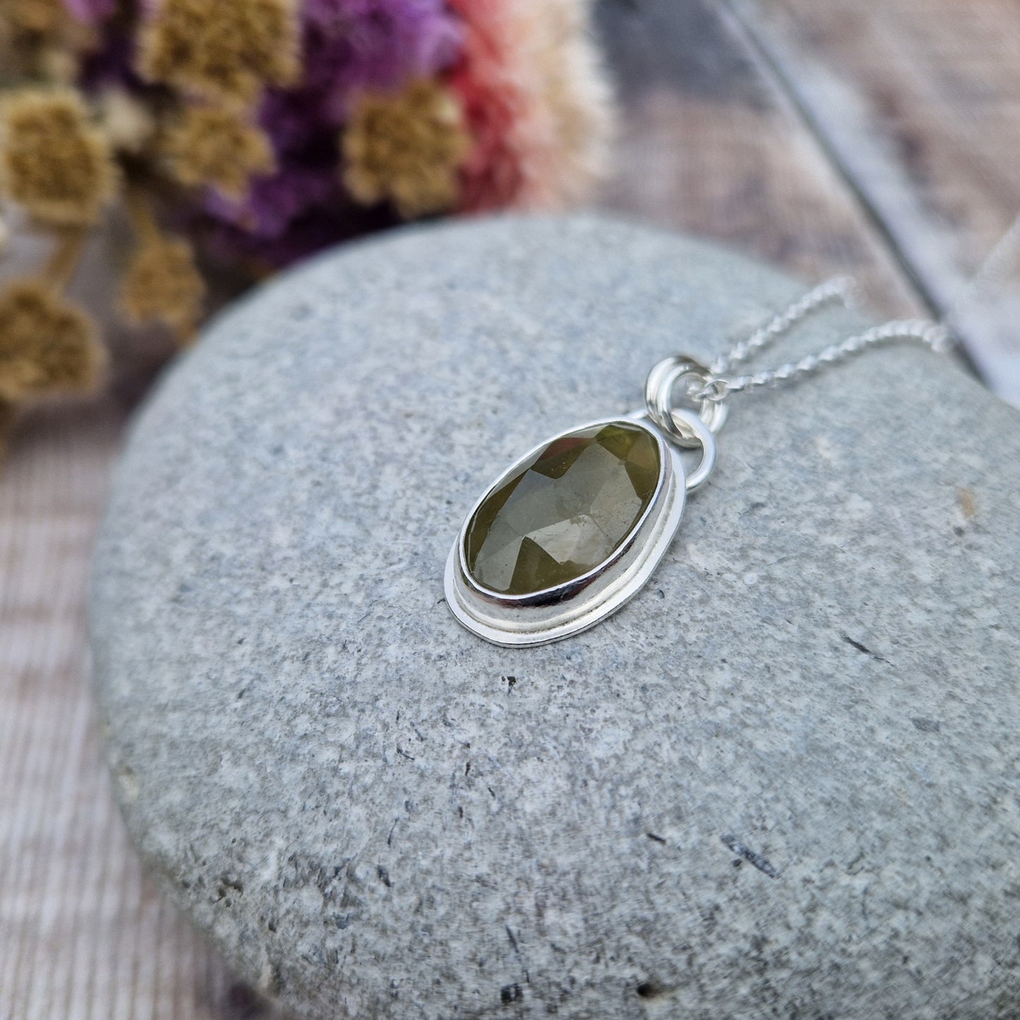 Green sapphire oval gemstone with diamond cut texture, set in simple silver bezel, attached to silver chain via two small circle links.