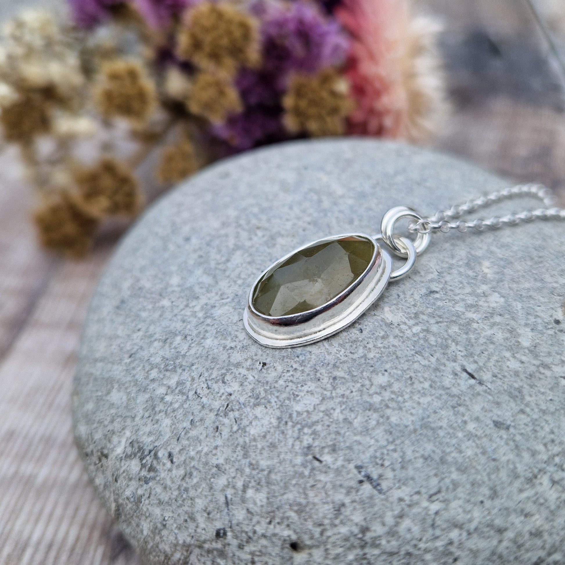 Green sapphire oval gemstone with diamond cut texture, set in simple silver bezel, attached to silver chain via two small circle links.