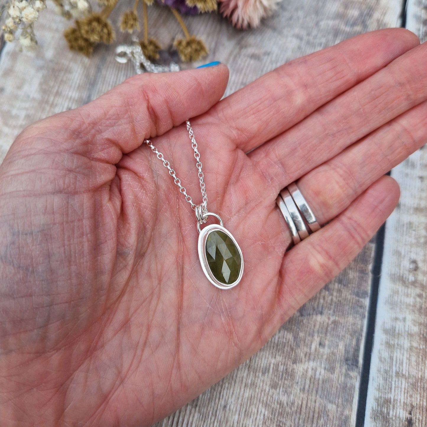 Green sapphire oval gemstone with diamond cut texture, set in simple silver bezel, attached to silver chain via two small circle links.