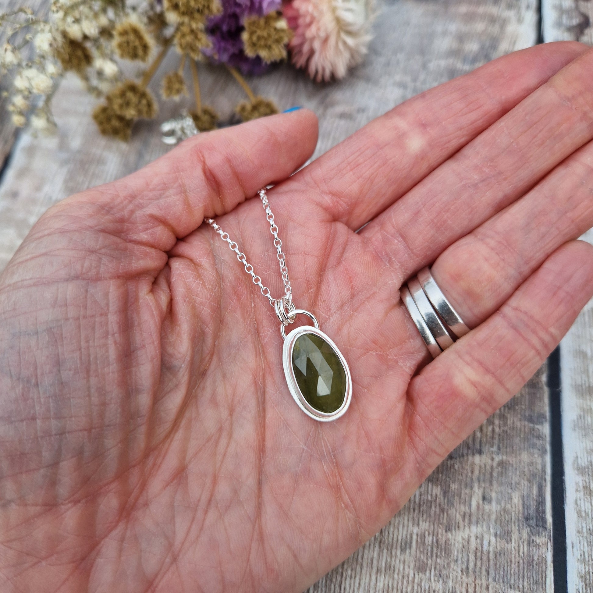 Green sapphire oval gemstone with diamond cut texture, set in simple silver bezel, attached to silver chain via two small circle links.